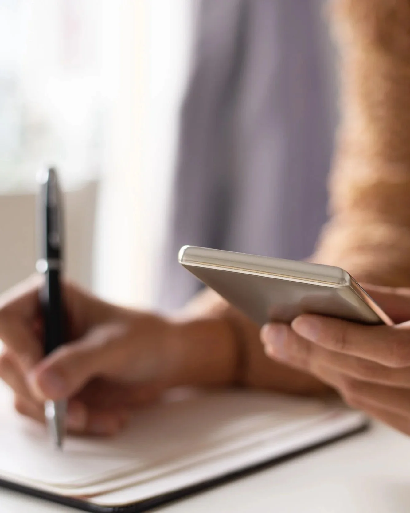 Persona escribiendo en un cuaderno con una mano y usando un teléfono móvil con la otra.