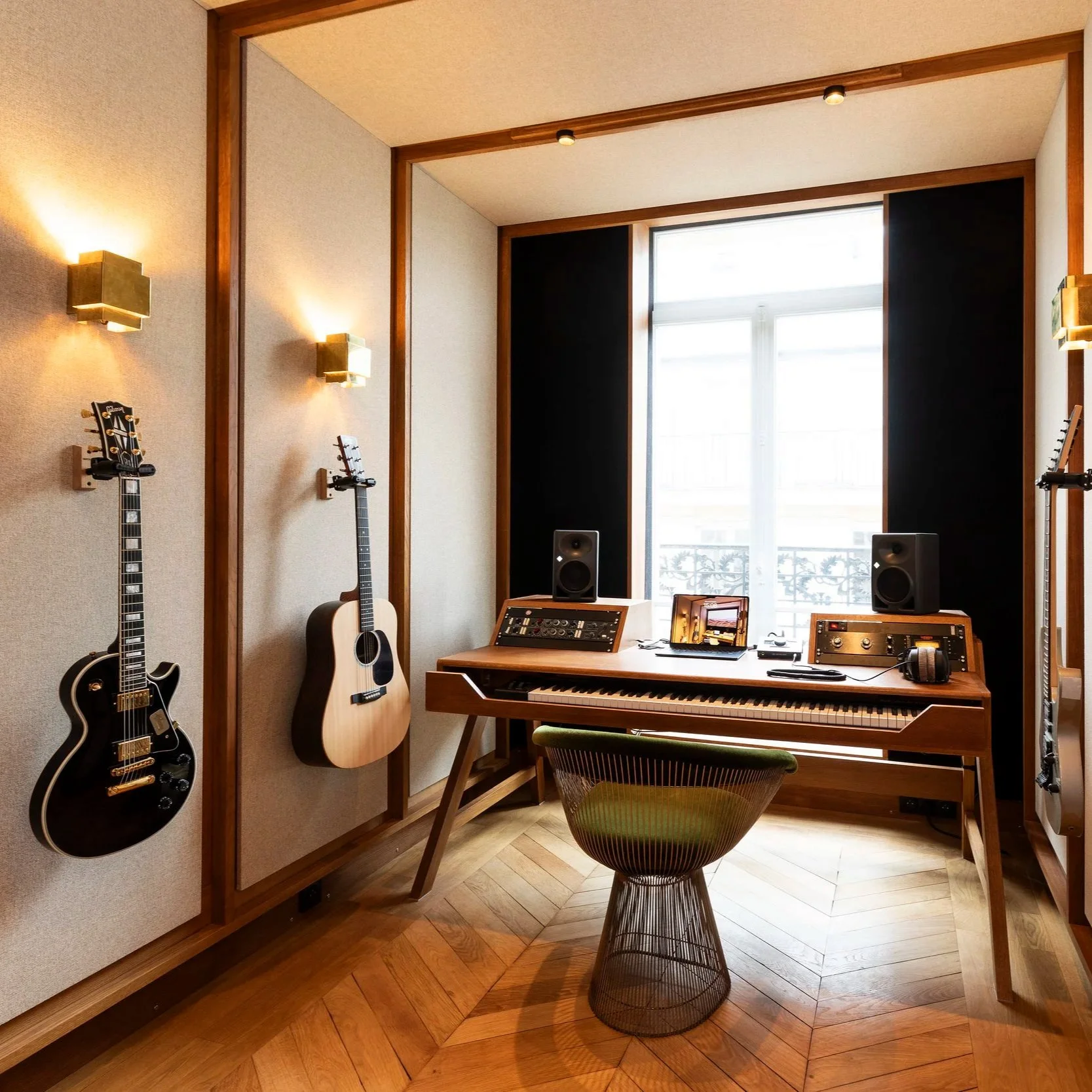 Studio d'enregistrement avec guitare électrique et acoustique accrochées au mur, un clavier, deux haut-parleurs, et divers équipements de musique sur un bureau en bois, dans une pièce lumineuse avec un grand fenêtre.