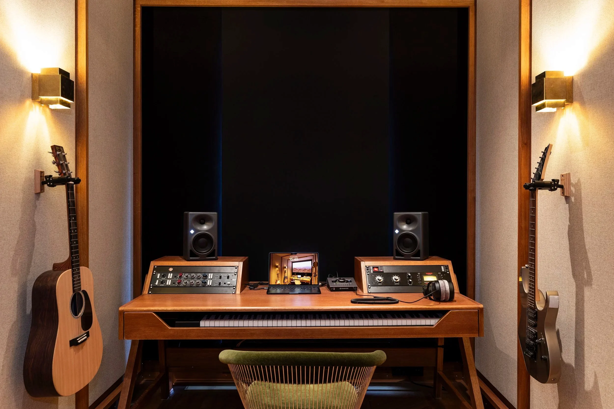 Studio d'enregistrement avec un piano, deux haut-parleurs, une guitare acoustique suspendue à gauche, une guitare électrique suspendue à droite, un ordinateur, un casque audio, et un micro dans un environnement avec des murs en tissu et un éclairage 