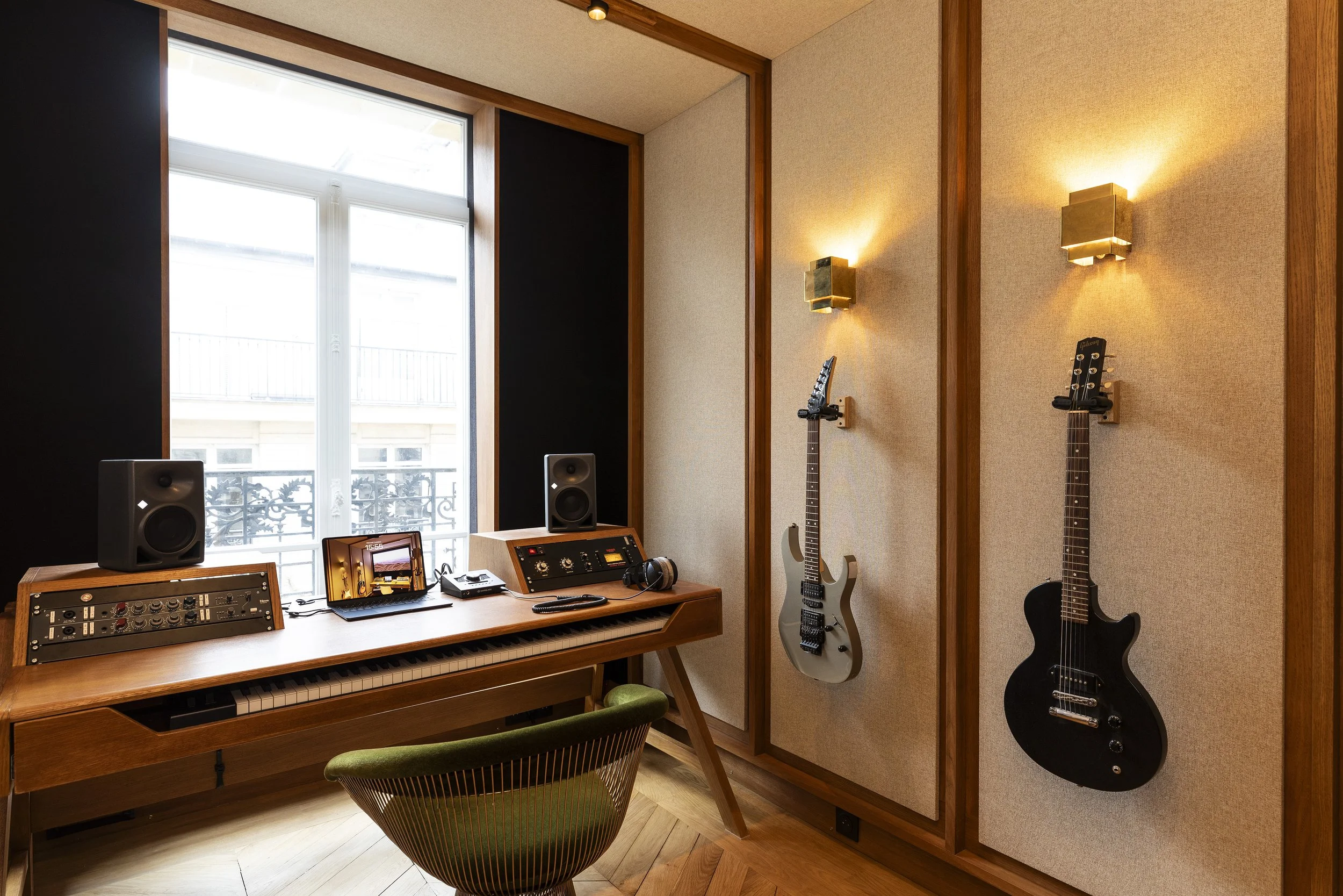 Studio d'enregistrement avec clavier, deux haut-parleurs, deux guitares fixées au mur, et un fauteuil vert.