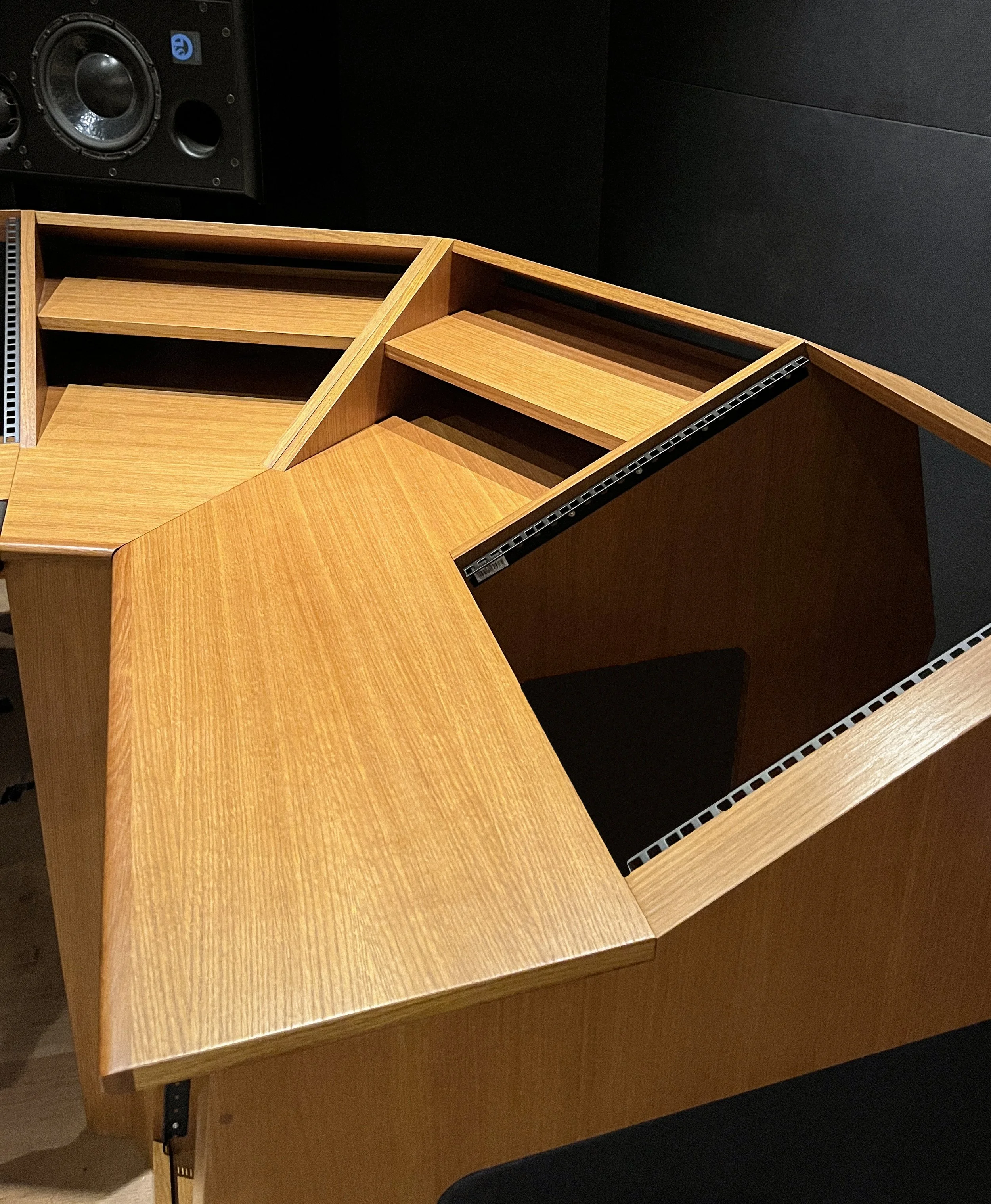 Meuble en bois avec compartiments de rangement et un plan de travail inclinable, dans une pièce sombre, avec une enceinte audio en arrière-plan.