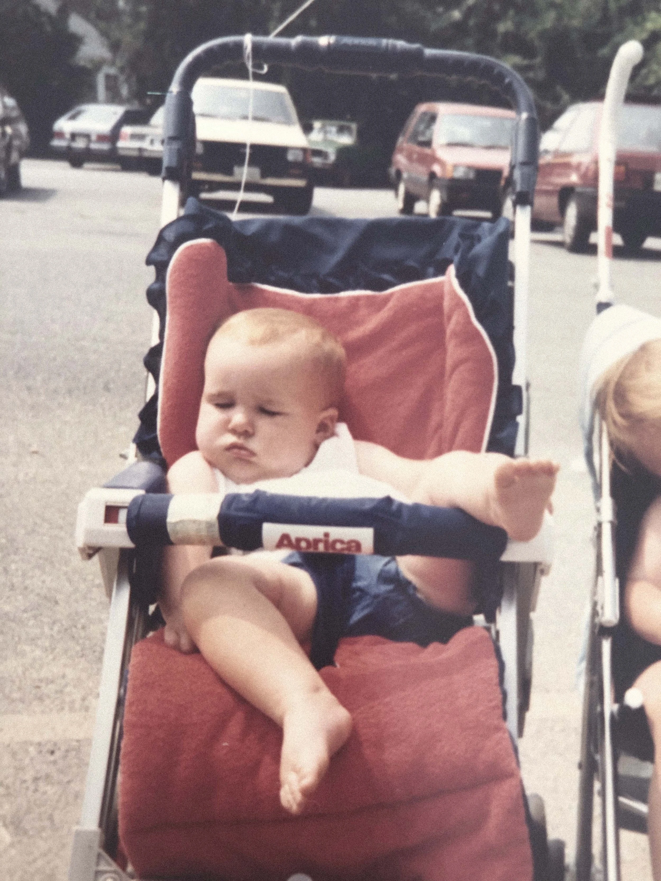 A baby sitting in a red and navy stroller outdoors in a parking lot with several cars in the background.
