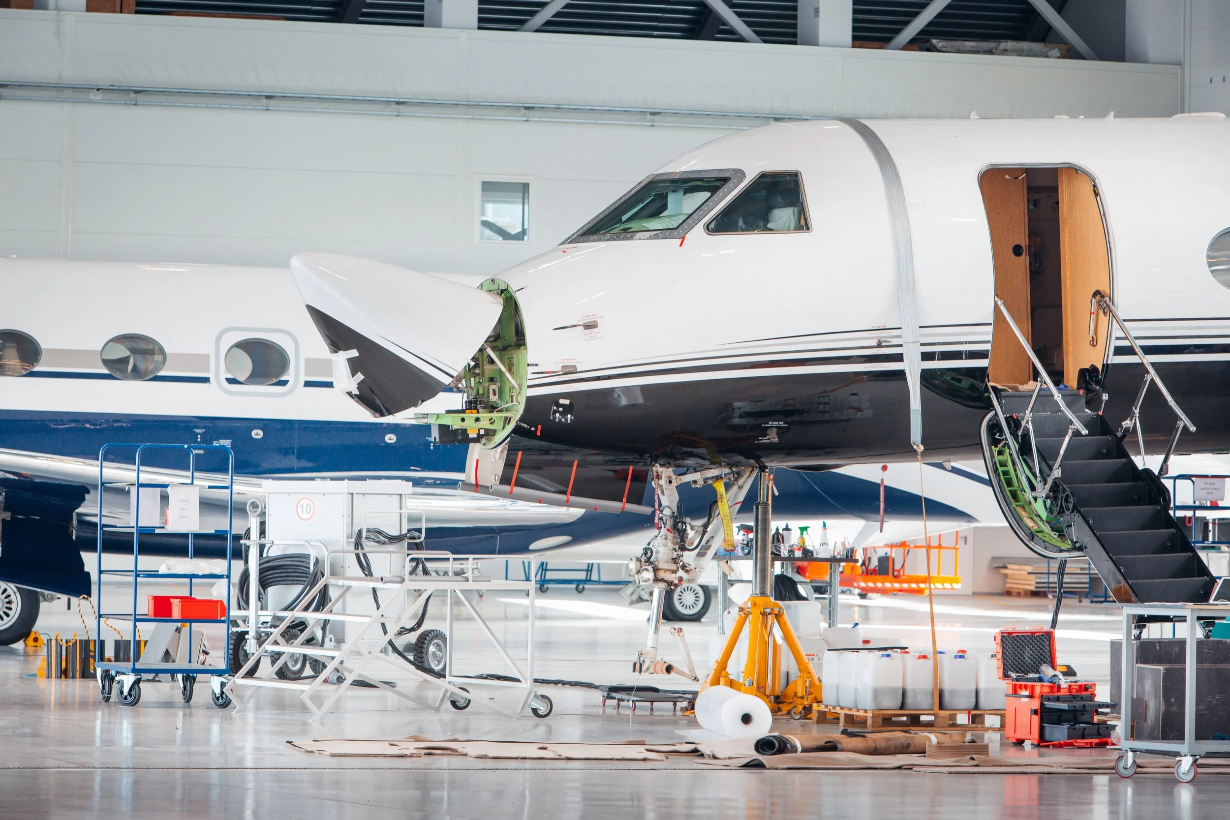Inside an airplane  maintenance facility, an aircraft is undergoing avionics repairs and upgrades with various tools and equipment around it.