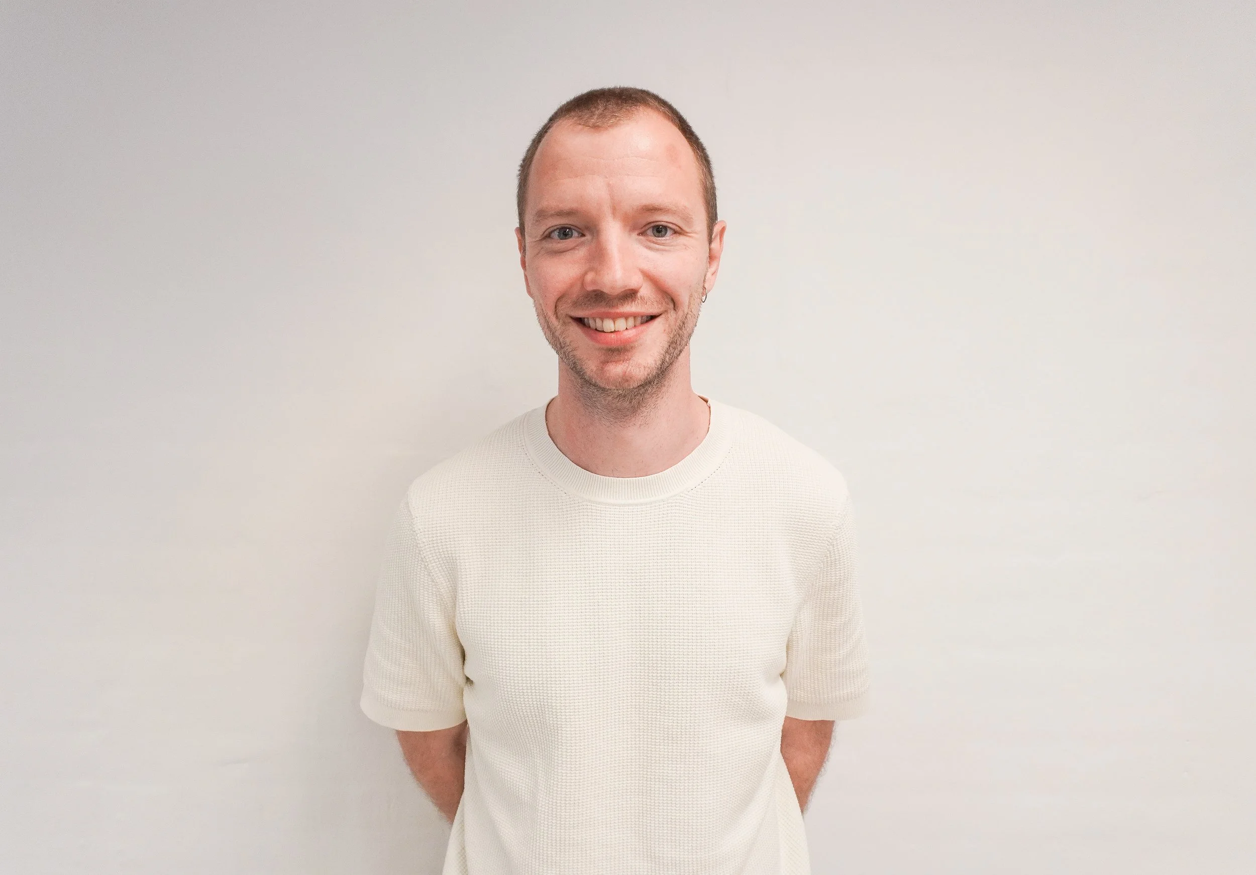 A smiling man with short hair and a beard, wearing a cream-colored sweater, standing against a plain white wall.