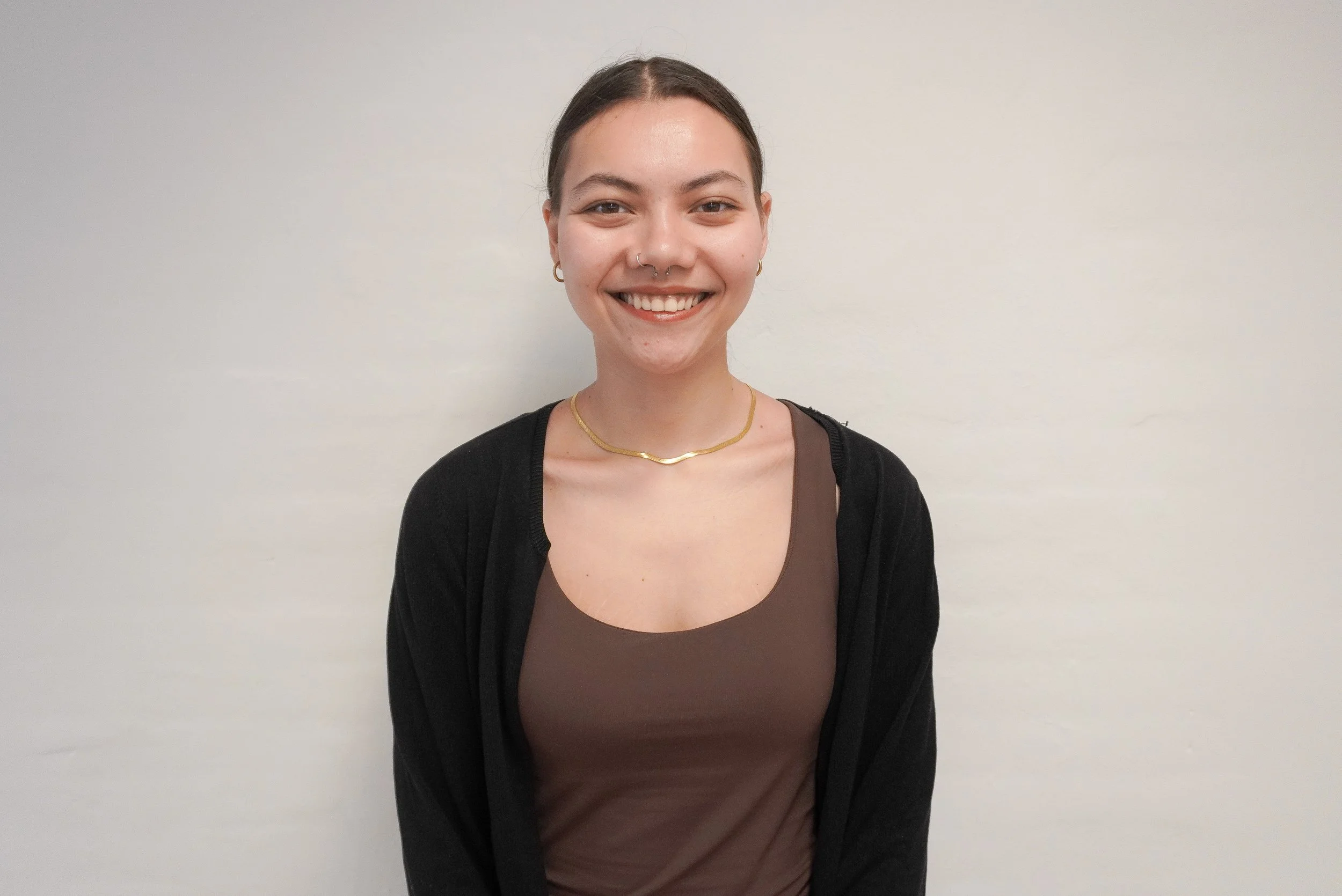 A young woman with dark hair, smiling, wearing a black cardigan, a brown top, gold jewelry, standing against a plain white wall.