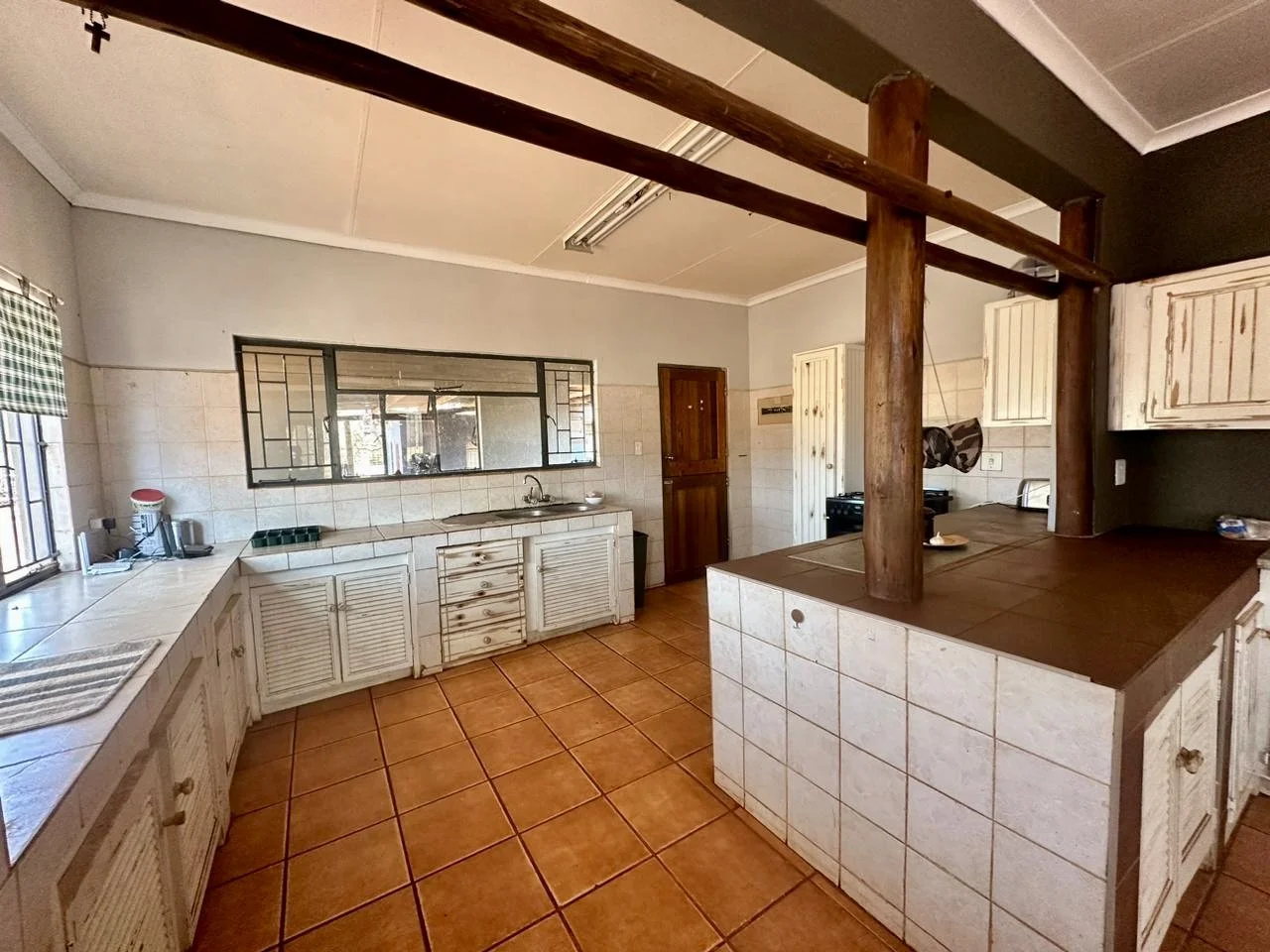 A kitchen with white cabinets, tiled countertops, and an orange tiled floor. It features a window with checkered curtains, a sink, and a wooden breakfast bar with a support beam.