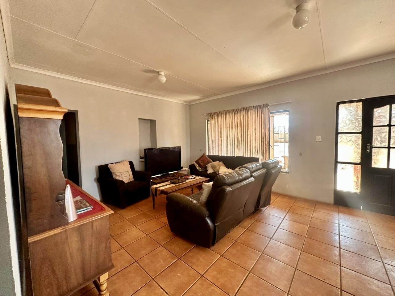 Living room with leather sofas, a wooden coffee table, a TV, an armchair, a side table, and windows with curtains, with tiled floor and a front door.