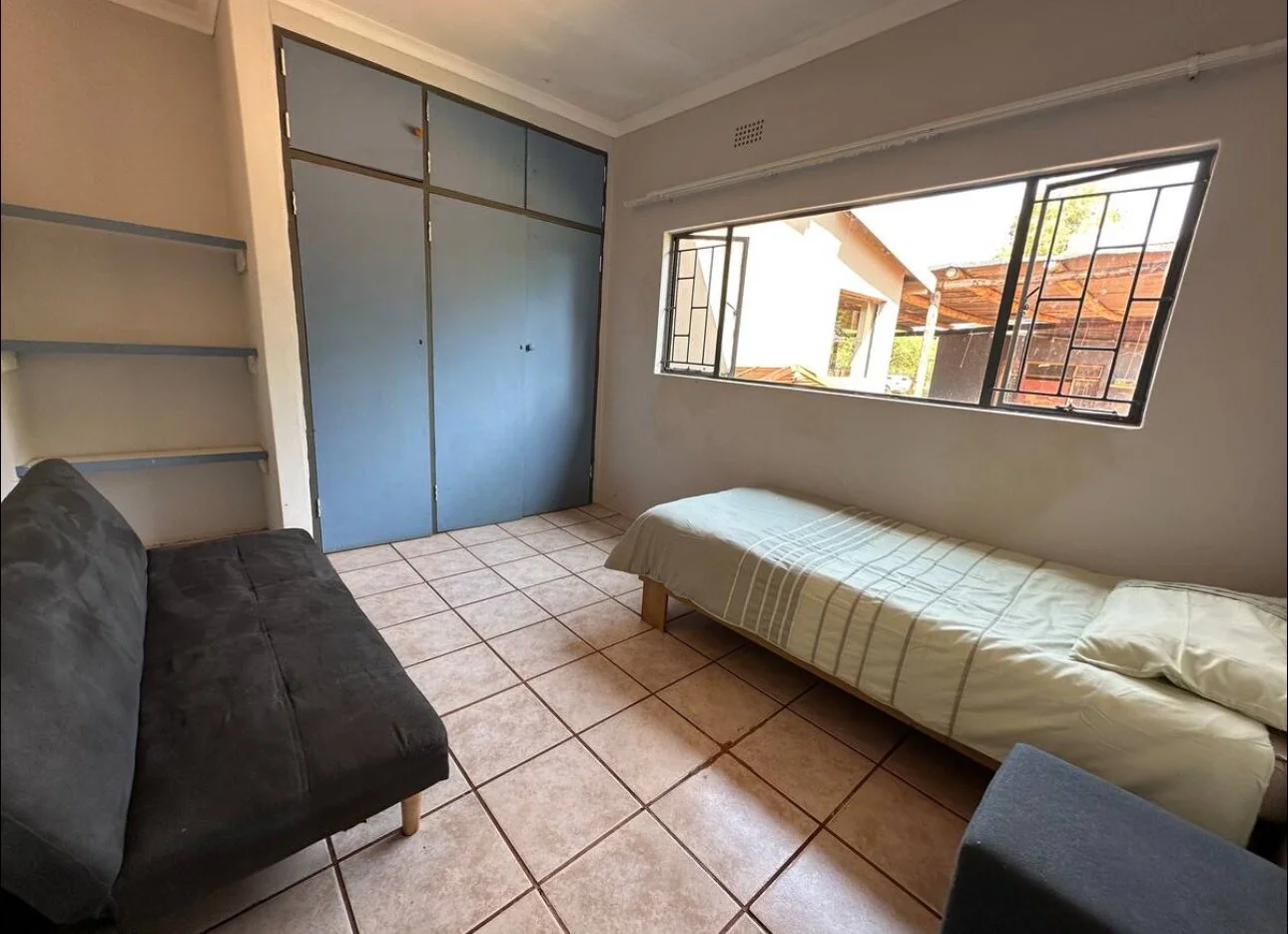 A small room with a black couch, a single bed with light-colored bedding, tiled floor, large window with metal bars, and a closed blue closet door.