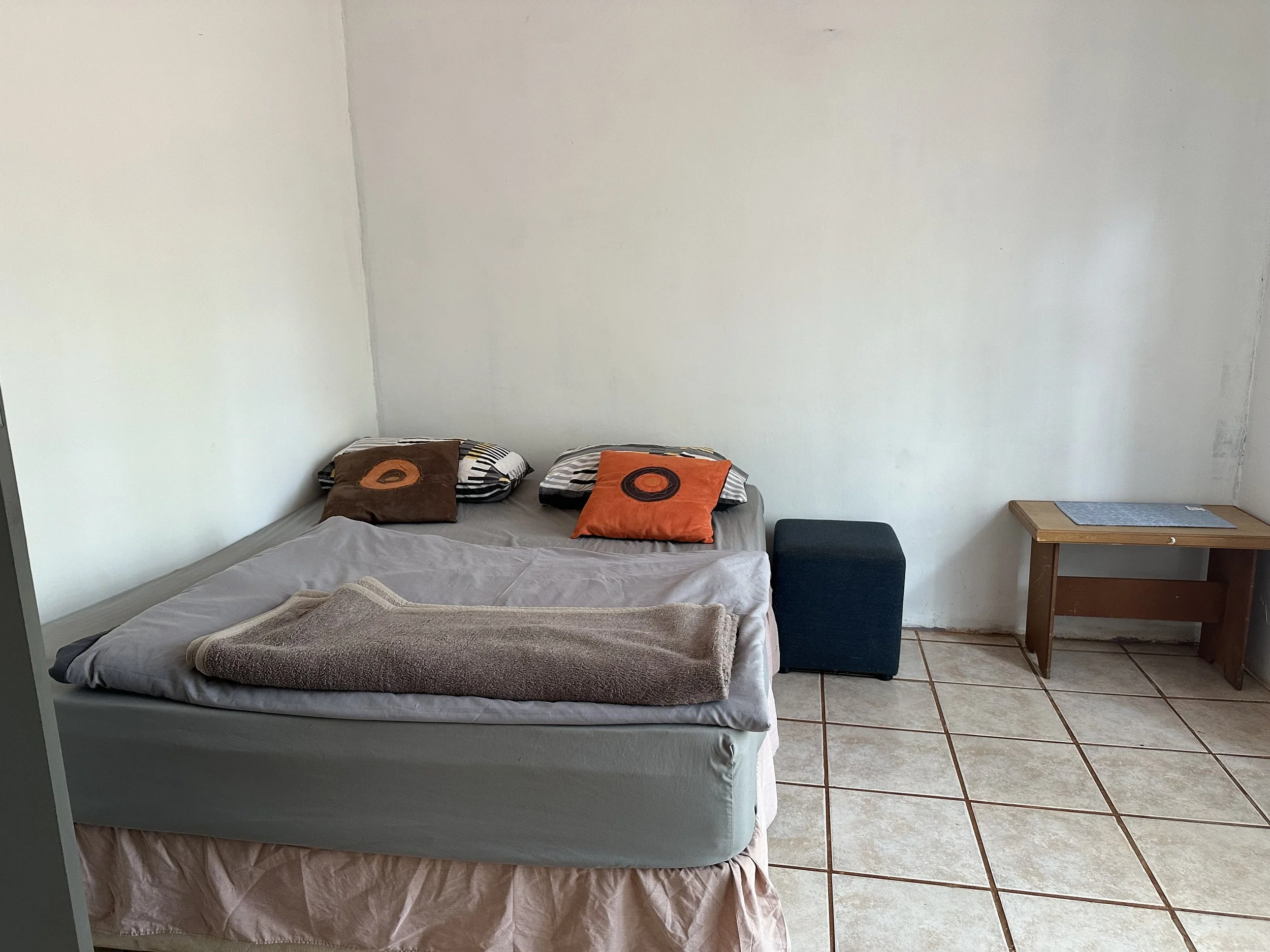 A bedroom with a bed covered in a gray sheet, three pillows with orange and brown pillowcases, a beige towel on the bed, a small wooden side table with gray mats on top, a black ottoman, and white walls with a tiled floor.
