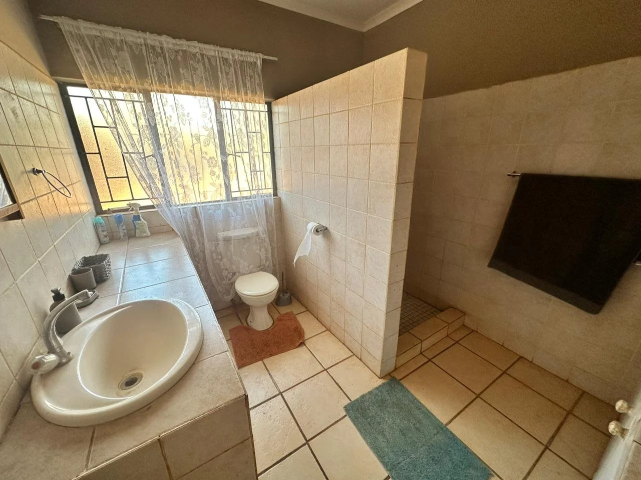A small bathroom with beige tiled walls and floor, a white sink with a faucet on the left, toiletries on the counter, a window with floral lace curtains letting in natural light, a toilet with a brown rug in front, a partial wall with a toilet paper 