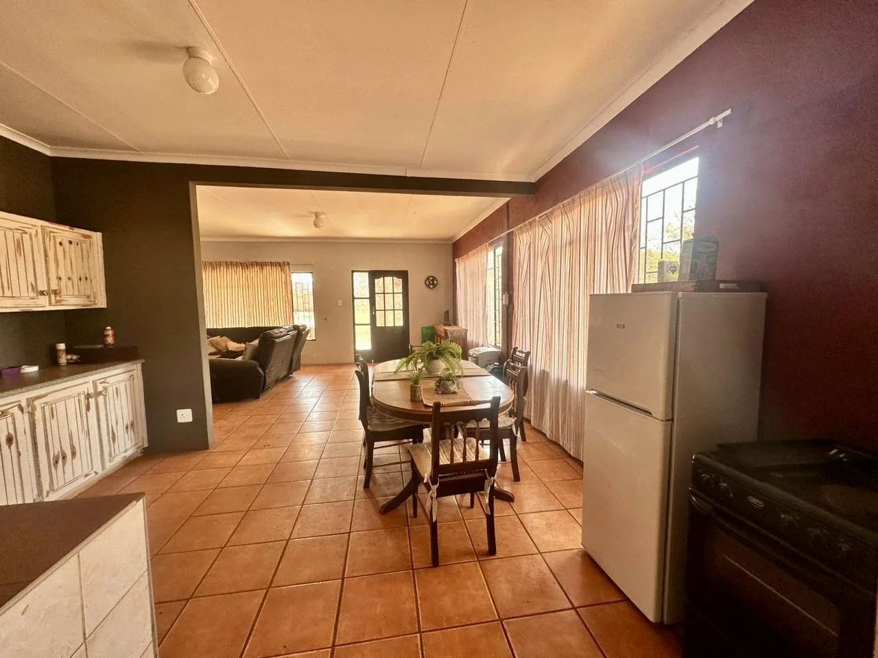 Interior of a living and dining room with terracotta tile flooring, a kitchen area on the right with a white refrigerator and stove, a round dining table with chairs, a black couch in the background, large windows with curtains, and a door leading ou