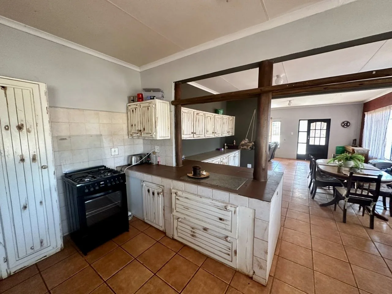 Open kitchen with white distressed cabinets, a black stove, tiled floor, and a wooden breakfast bar with a coffee cup on a placemat. Adjacent to the kitchen is a dining area with a round table, chairs, and a sliding glass door with curtains.
