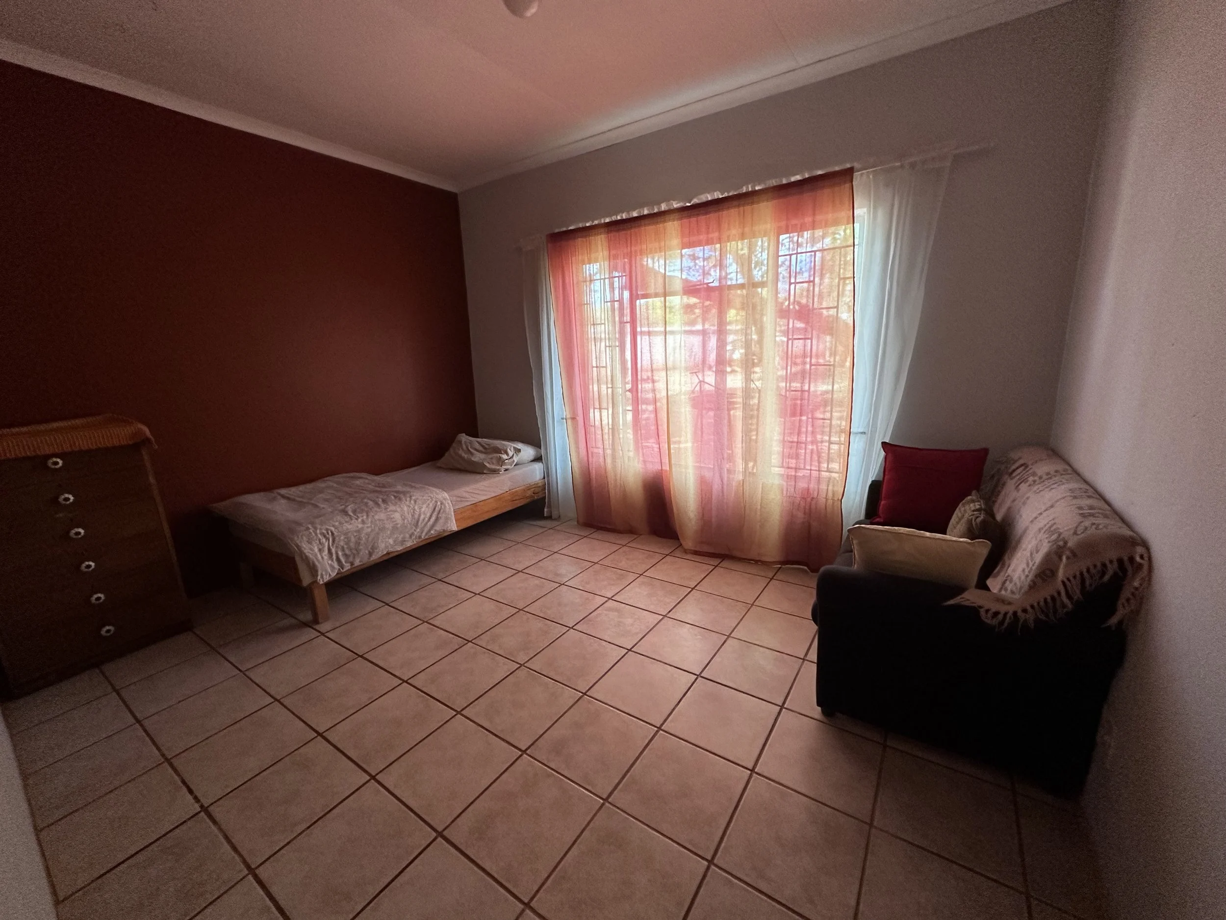 A room with tiled floor, a bed with pillows, a dresser, a window with red and white curtains, and a sofa with a blanket and pillows.