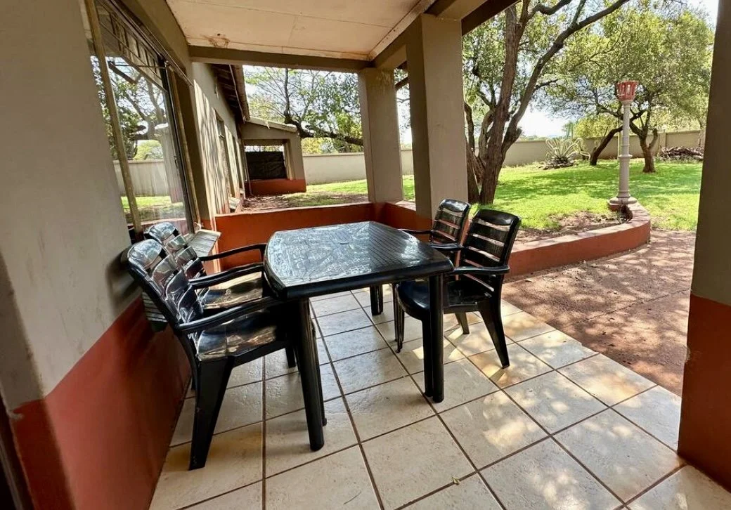An outdoor patio with a black plastic table and four matching chairs, surrounded by a garden with green grass, trees, and a lamppost.