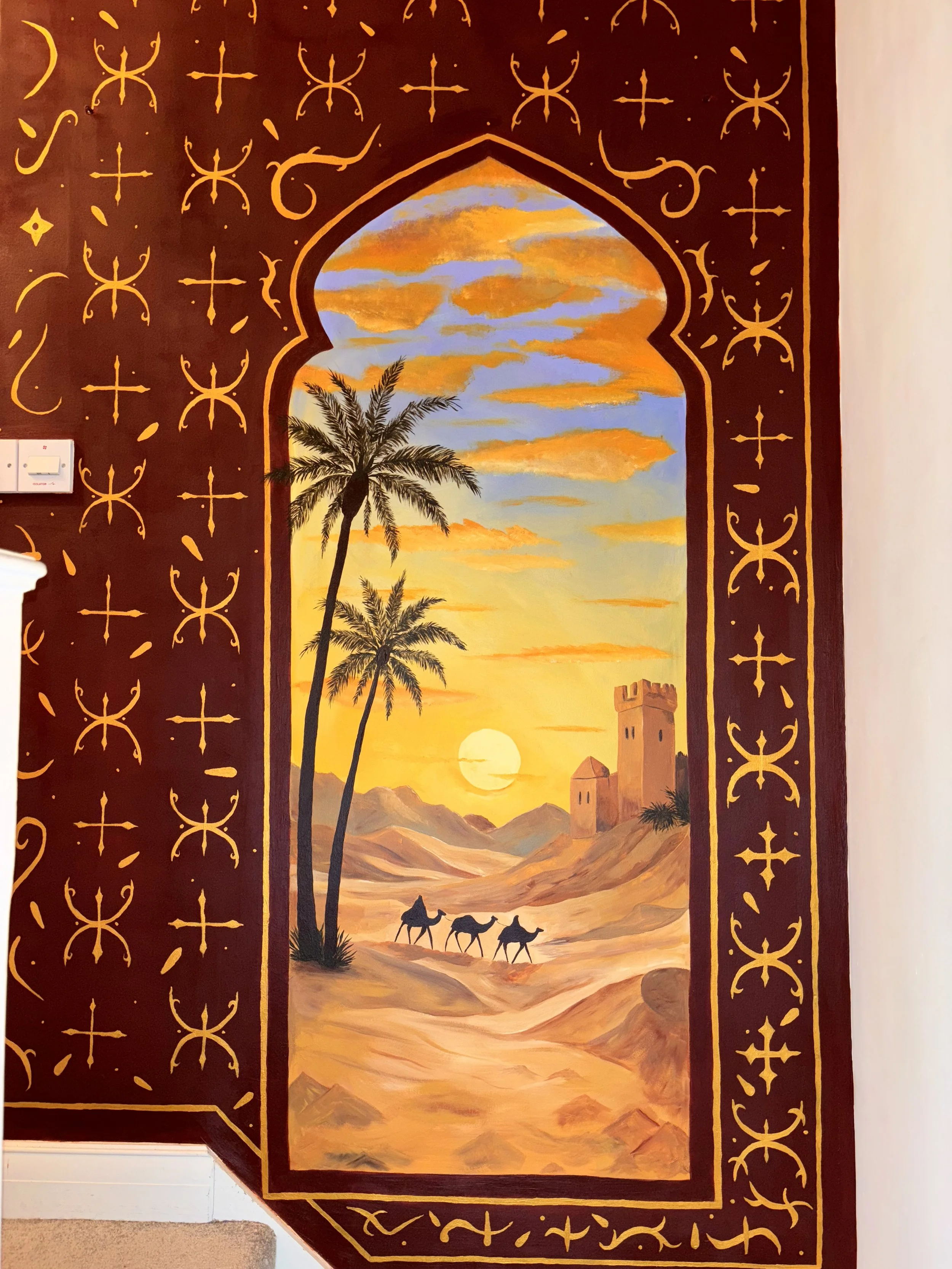bohemian internal mural hand painted in warm, rich colours. wall art framed in a deep red border with repeating gold geometric and symbolic patterns. arched frame, desert landscape at sunset. soft sand dunes. camel, palm trees Middle Eastern decor uk