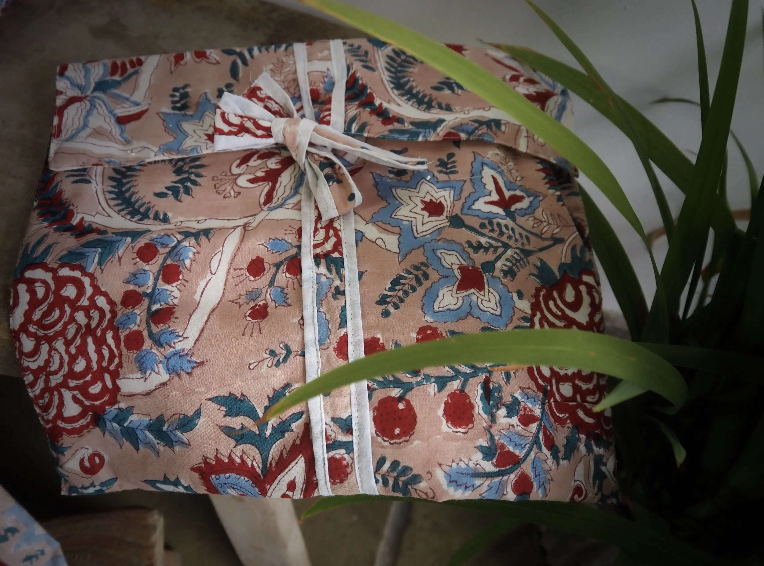 A neatly wrapped gift box with floral and fruit patterns in red, blue, and beige, tied with a white ribbon, resting on a surface next to green leafy plants.