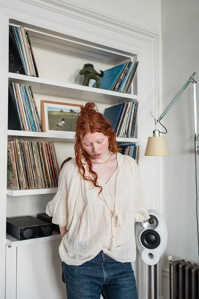 A woman with red hair standing in front of a white bookshelf filled with records and books, with a speaker and a lamp nearby.