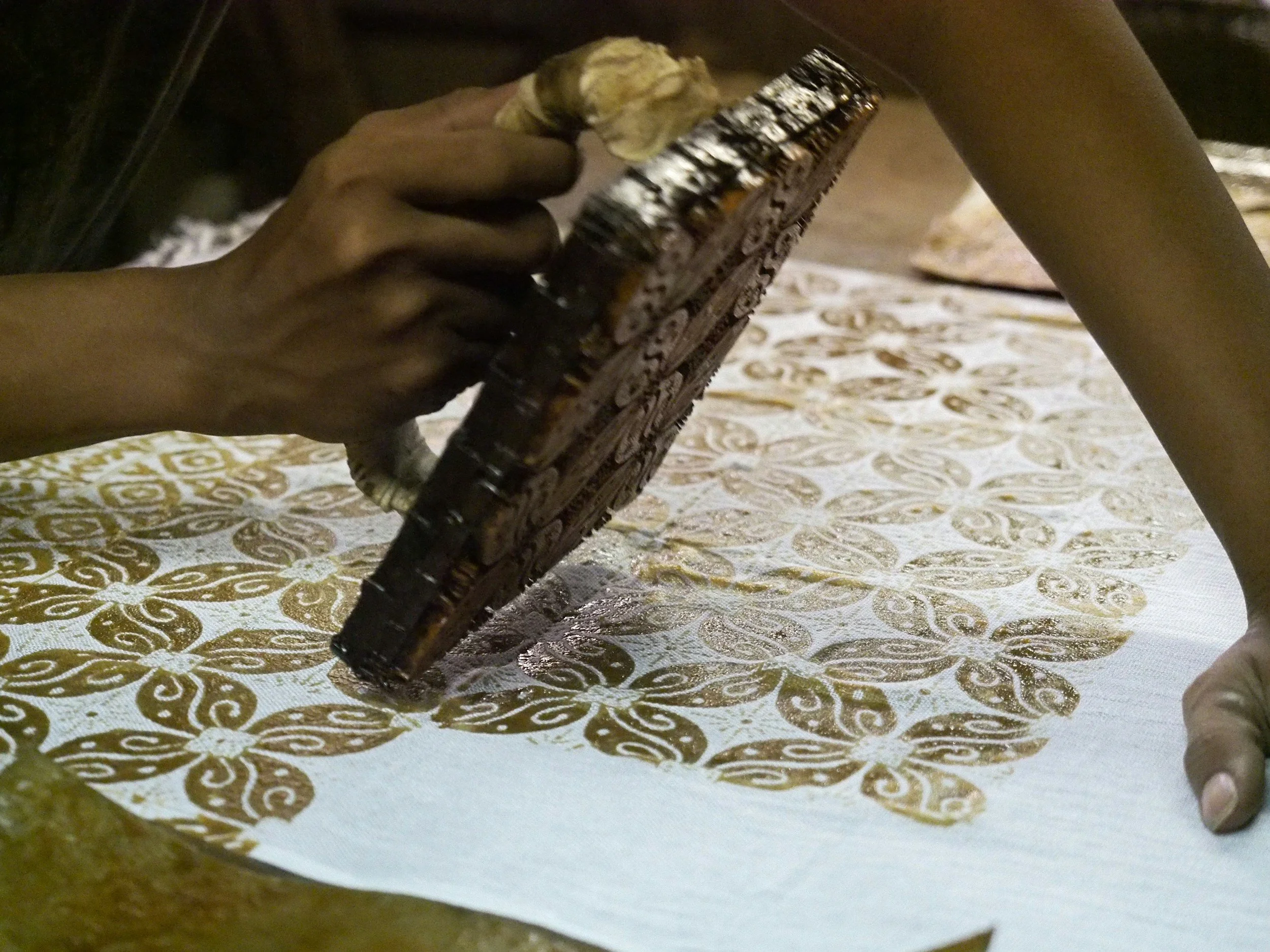 Person using a carved wooden block and a mallet to print a golden floral pattern on fabric.