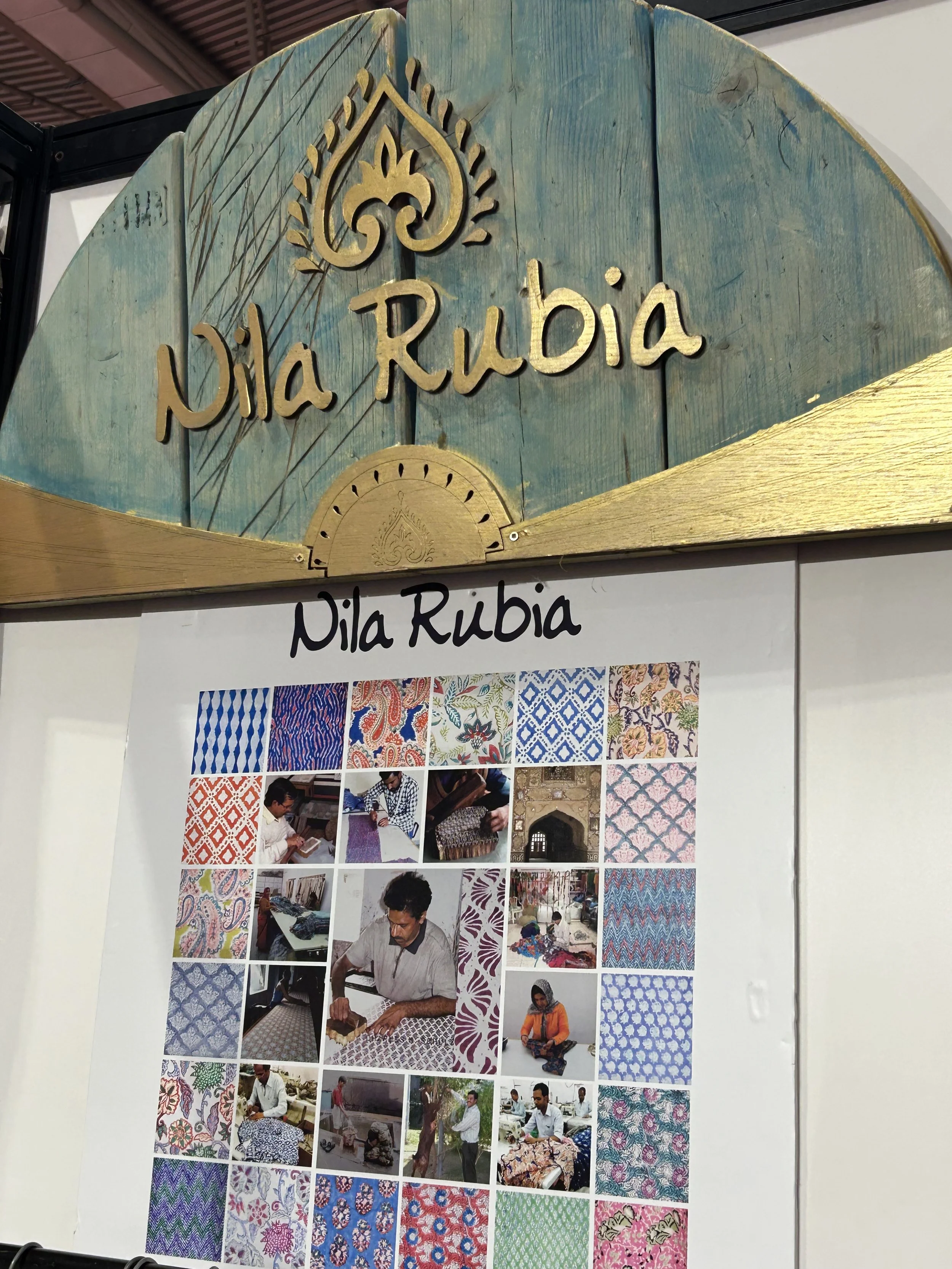 Photograph of a display board showing traditional fabric patterns and photos of artisans making fabric, with a wooden sign above reading 'Nila Rubia'.