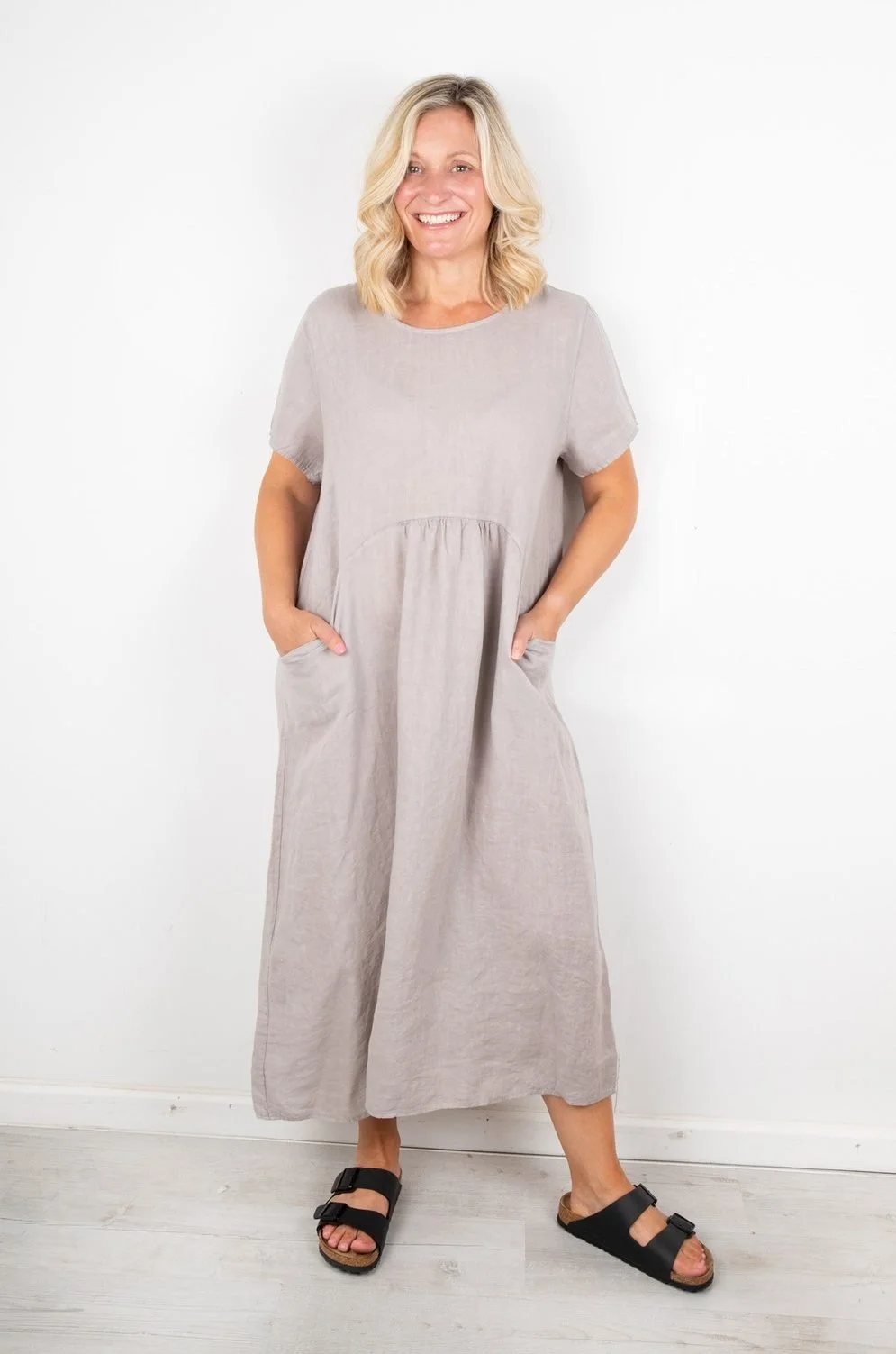 A woman with blonde hair is standing indoors against a white wall, smiling, wearing a light gray jumpsuit with short sleeves and black sandals.