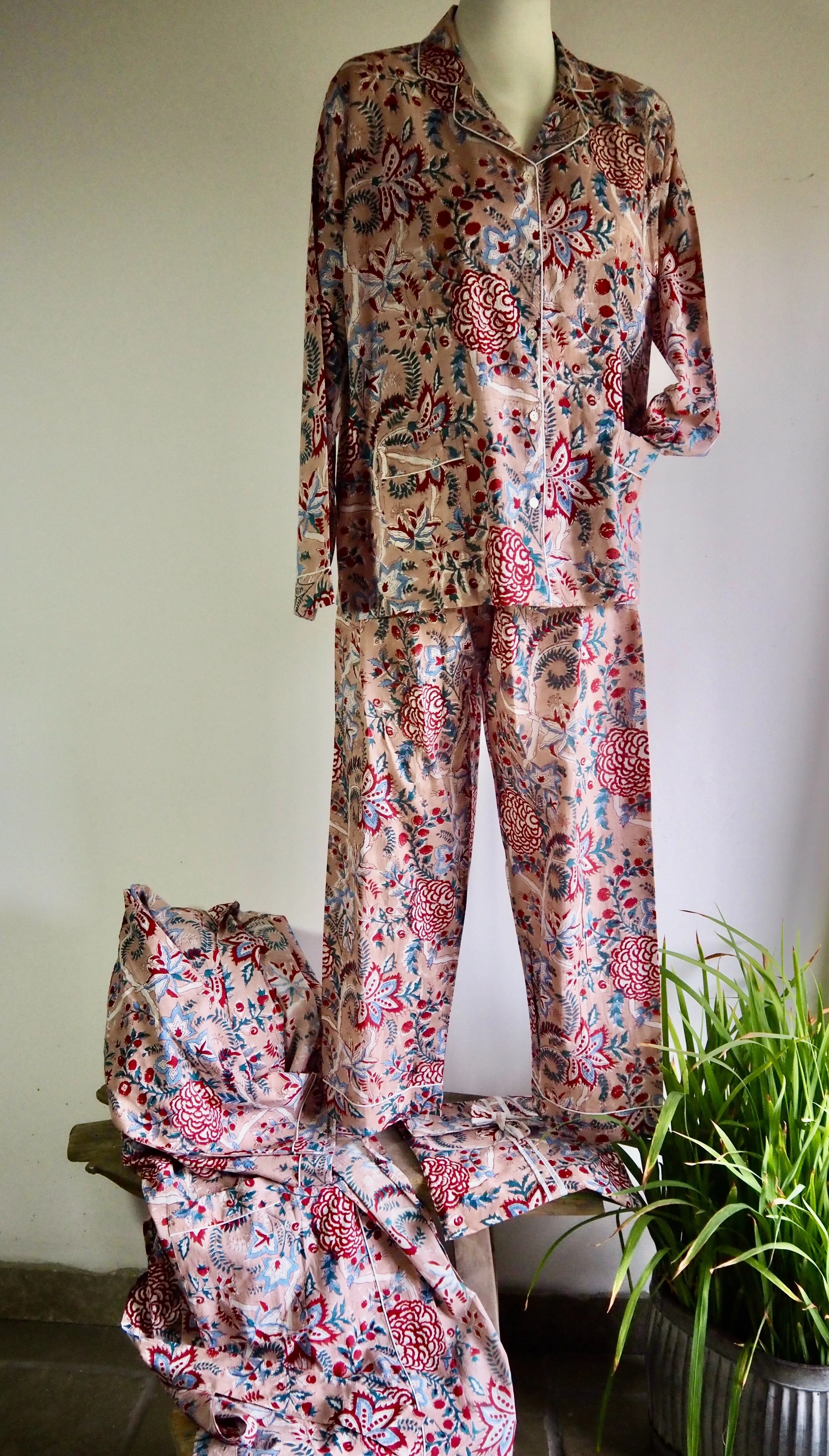A mannequin wearing a pink floral-patterned pajama set with a matching bag and pants, displayed on a wooden table with a potted green plant beside it.