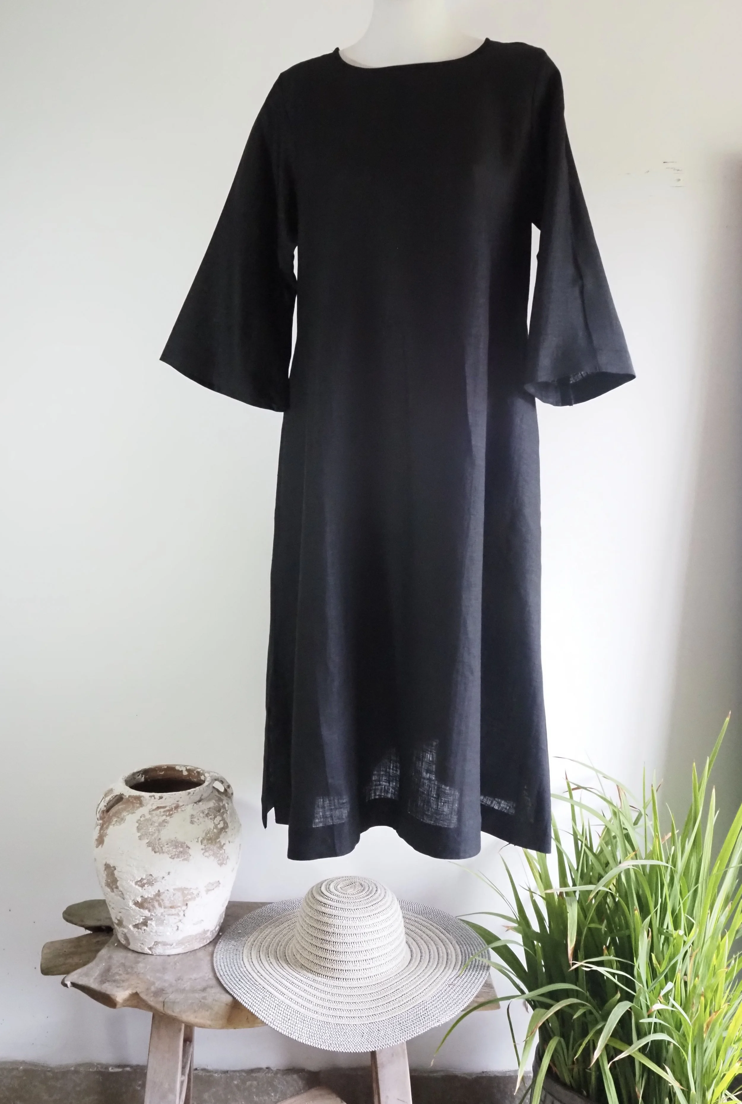 A black long dress with wide sleeves hanging on a white mannequin next to a small wooden table. On the table are a rustic ceramic vase, a light-colored wide-brimmed straw hat, and a potted green plant.