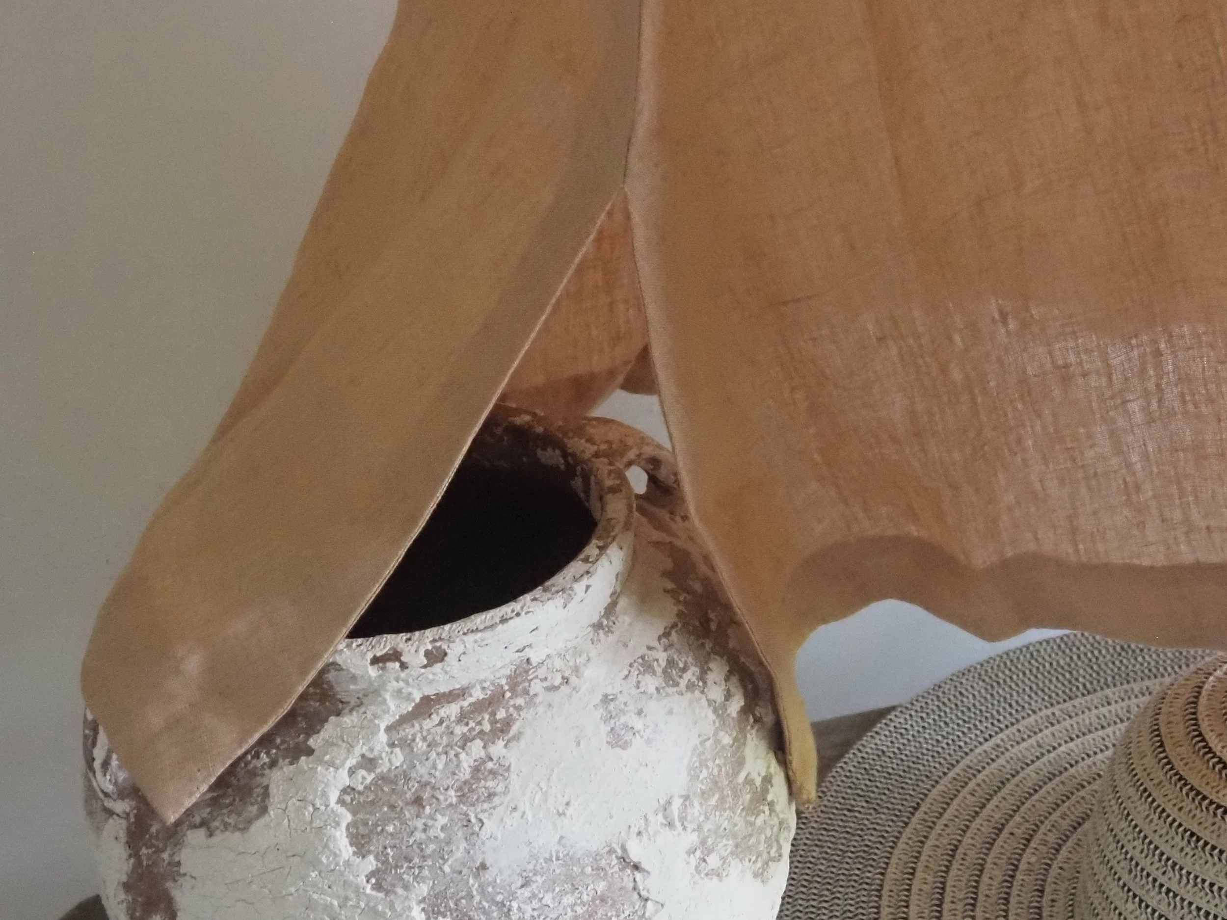 Close-up of a rustic, weathered white ceramic pitcher with dark stain spills, partially hidden behind a brown paper bag, on a surface with a woven circular placemat.