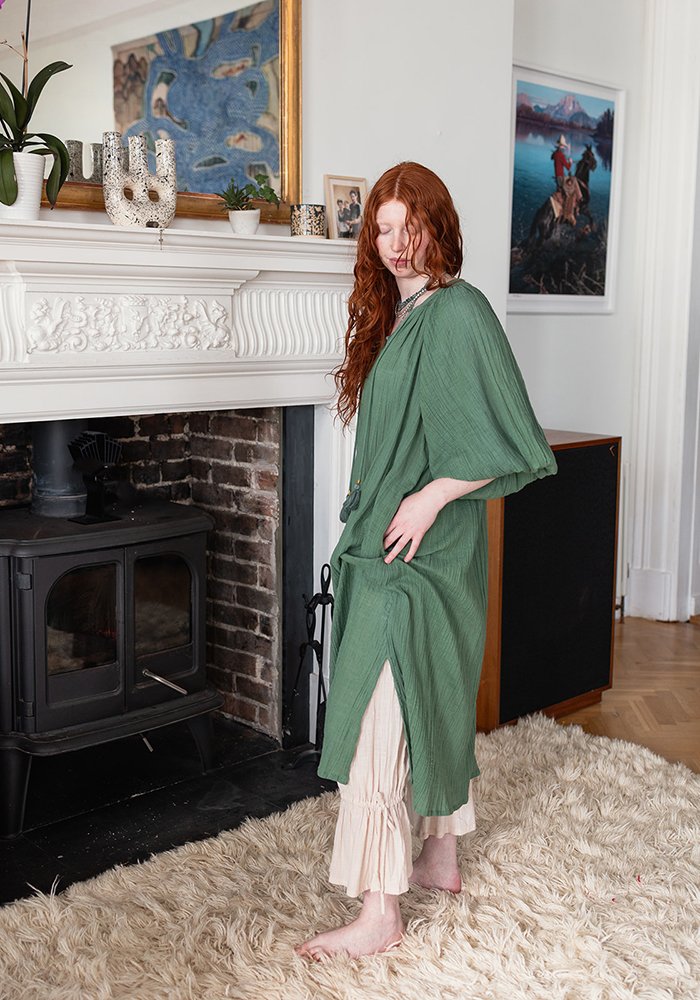 A woman with long red hair wearing a green robe and beige ruffled pants standing near a fireplace with a white mantel decorated with plants and art, in a cozy living room.