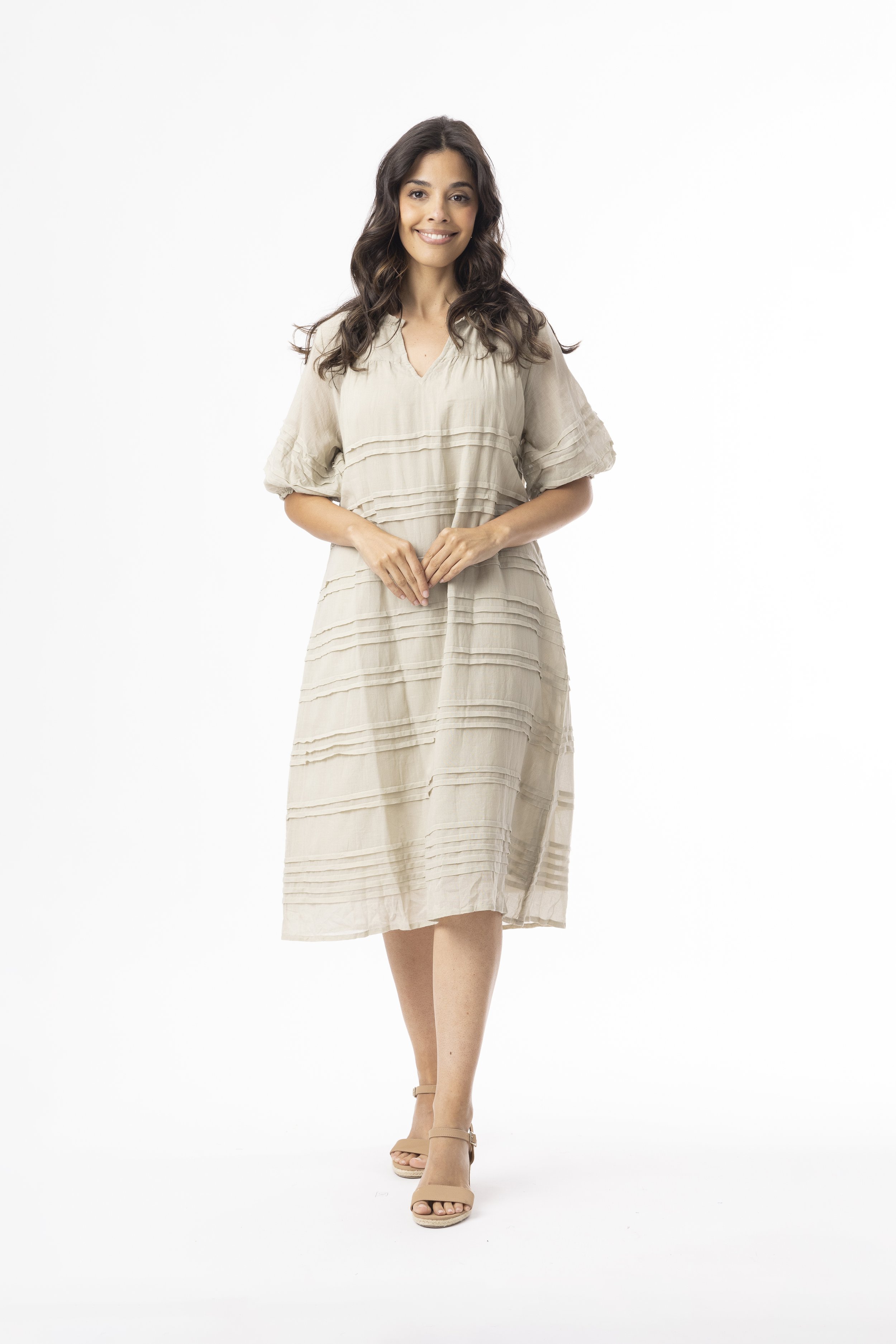 A woman with long dark curly hair, smiling, wearing a beige midi dress and nude high-heeled sandals, standing against a white background.