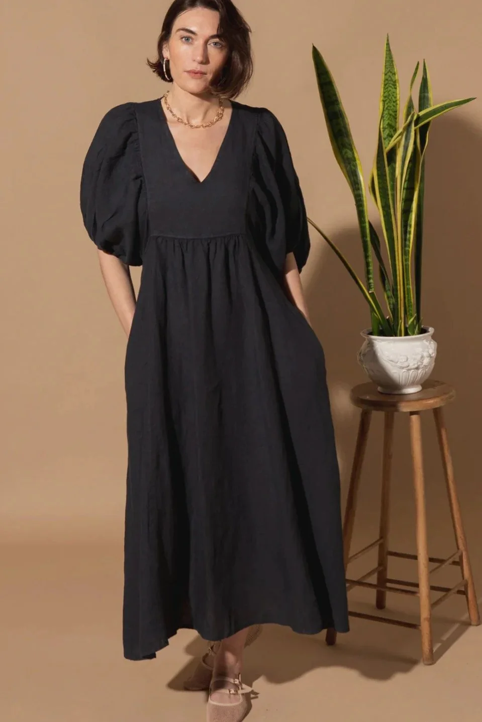 A woman in a black dress standing next to a potted plant on a small wooden table against a beige background.