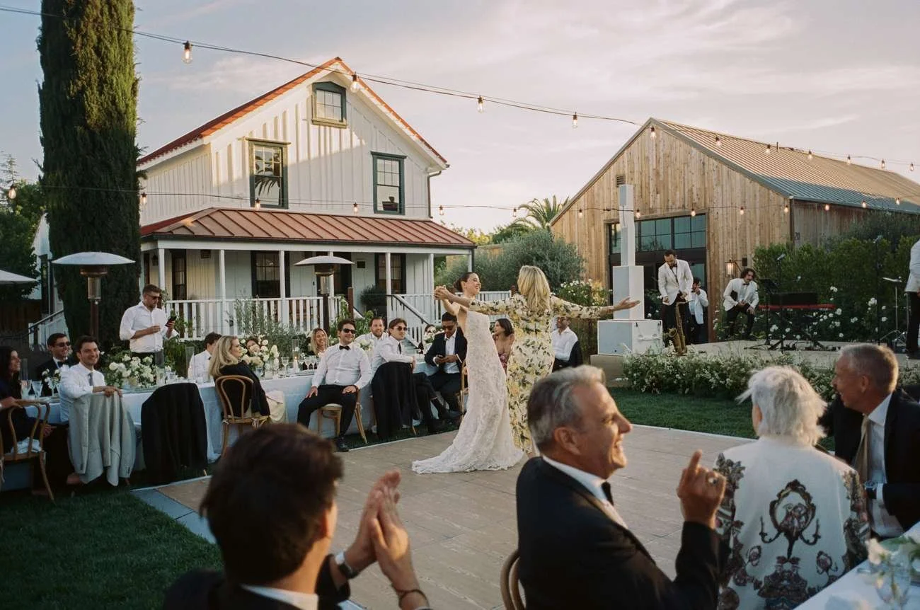 Outdoor wedding reception with guests watching brides dancing in front of house and barn.