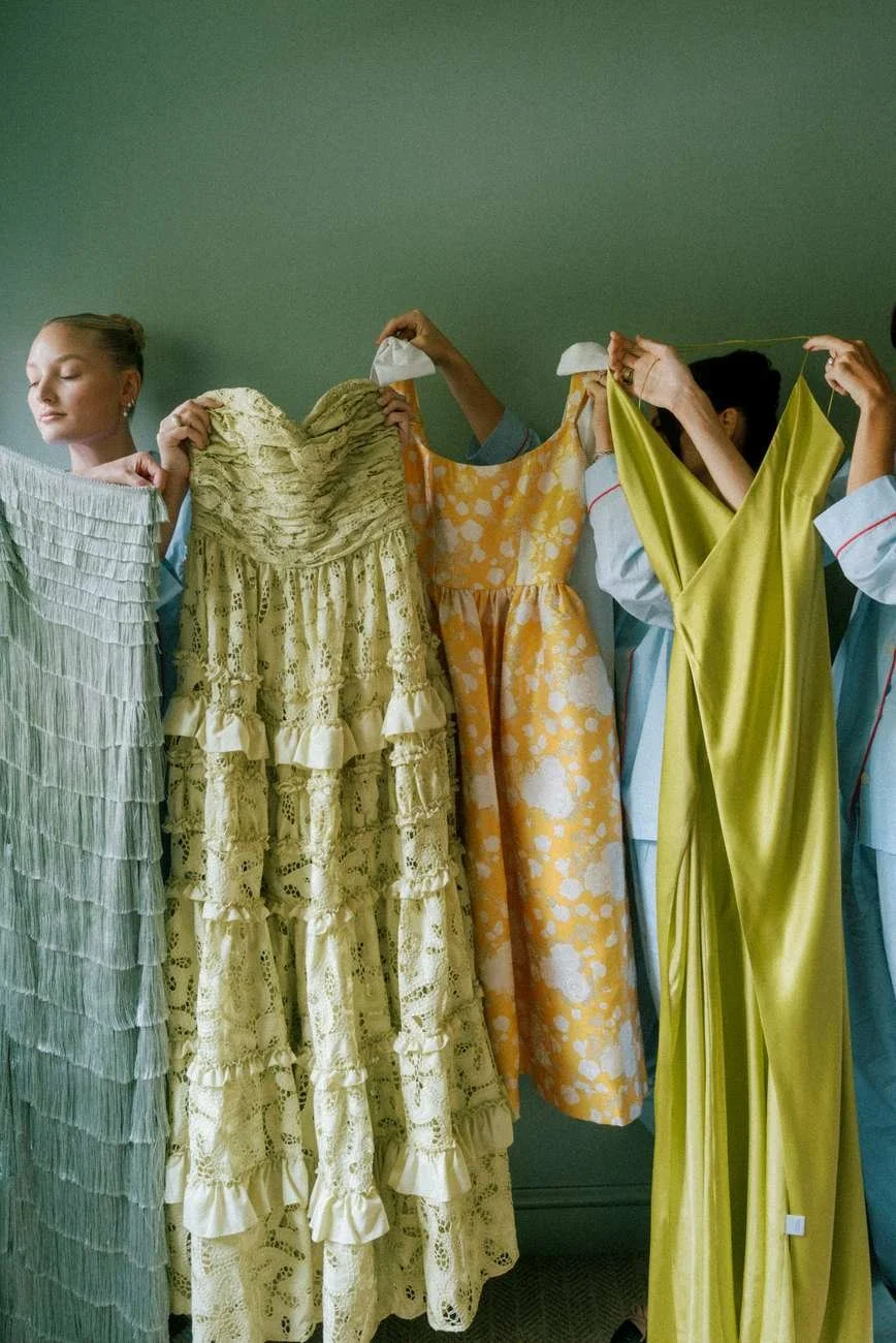 Women holding up colorful dresses against a green wall.