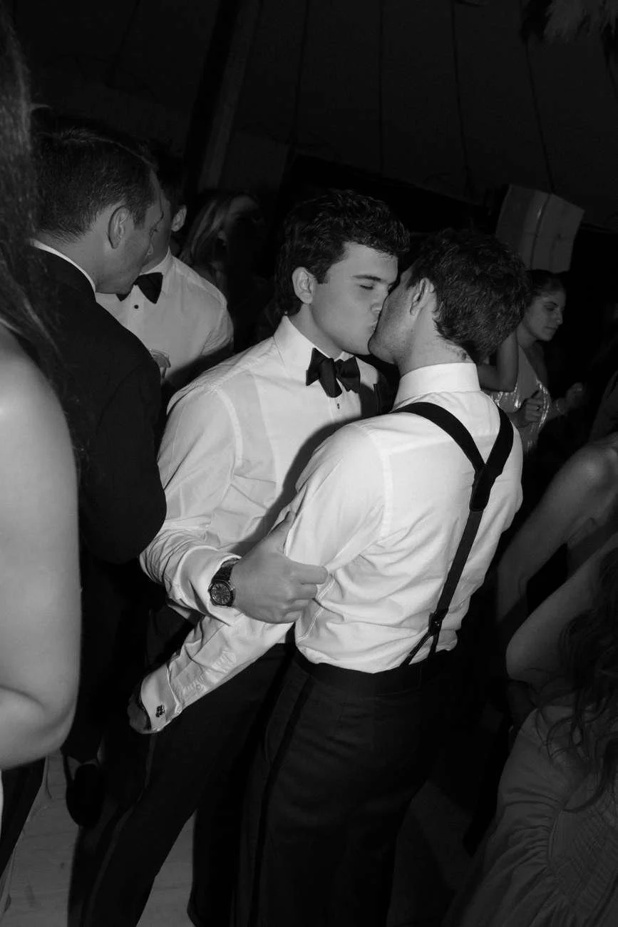 Two men in tuxedos kissing at a formal event.
