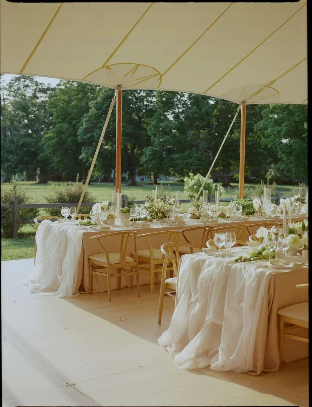 Elegant outdoor event setup with long tables draped in white cloth, decorated with floral centerpieces.