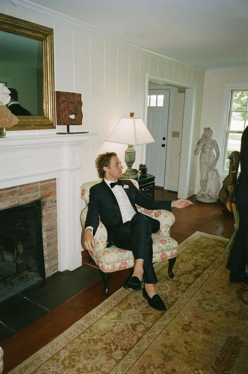 A man in a tuxedo sitting on a floral armchair next to a fireplace and lamp in a living room.