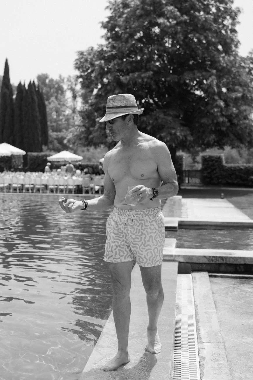 A shirtless man walking by a poolside on a sunny day, with trees and pool chairs in the background.