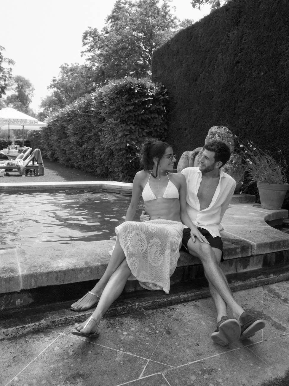 A couple sitting on the edge of a swimming pool, smiling and looking at each other.