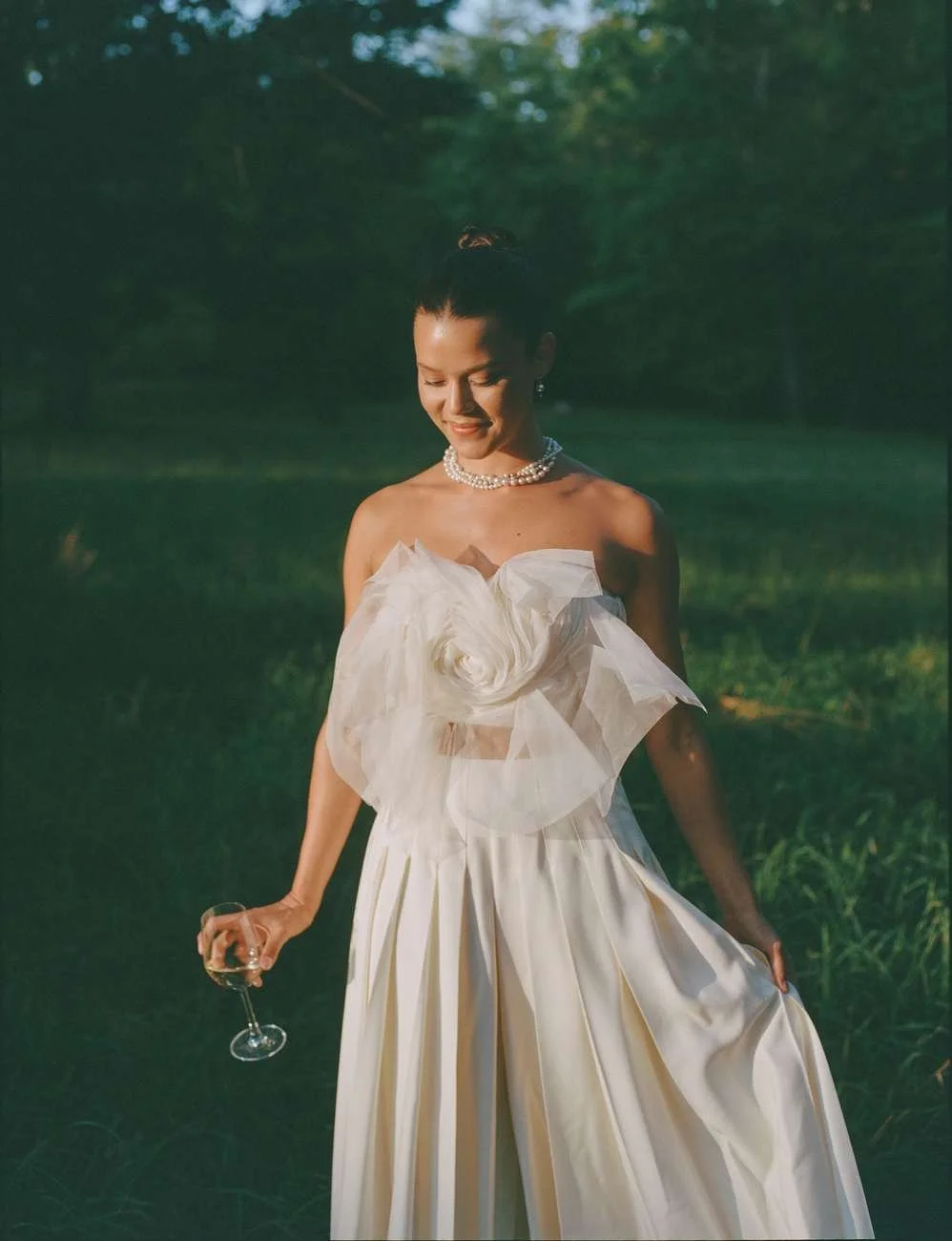 Woman in a white strapless dress holding a wine glass, smiling outdoors in a green park.