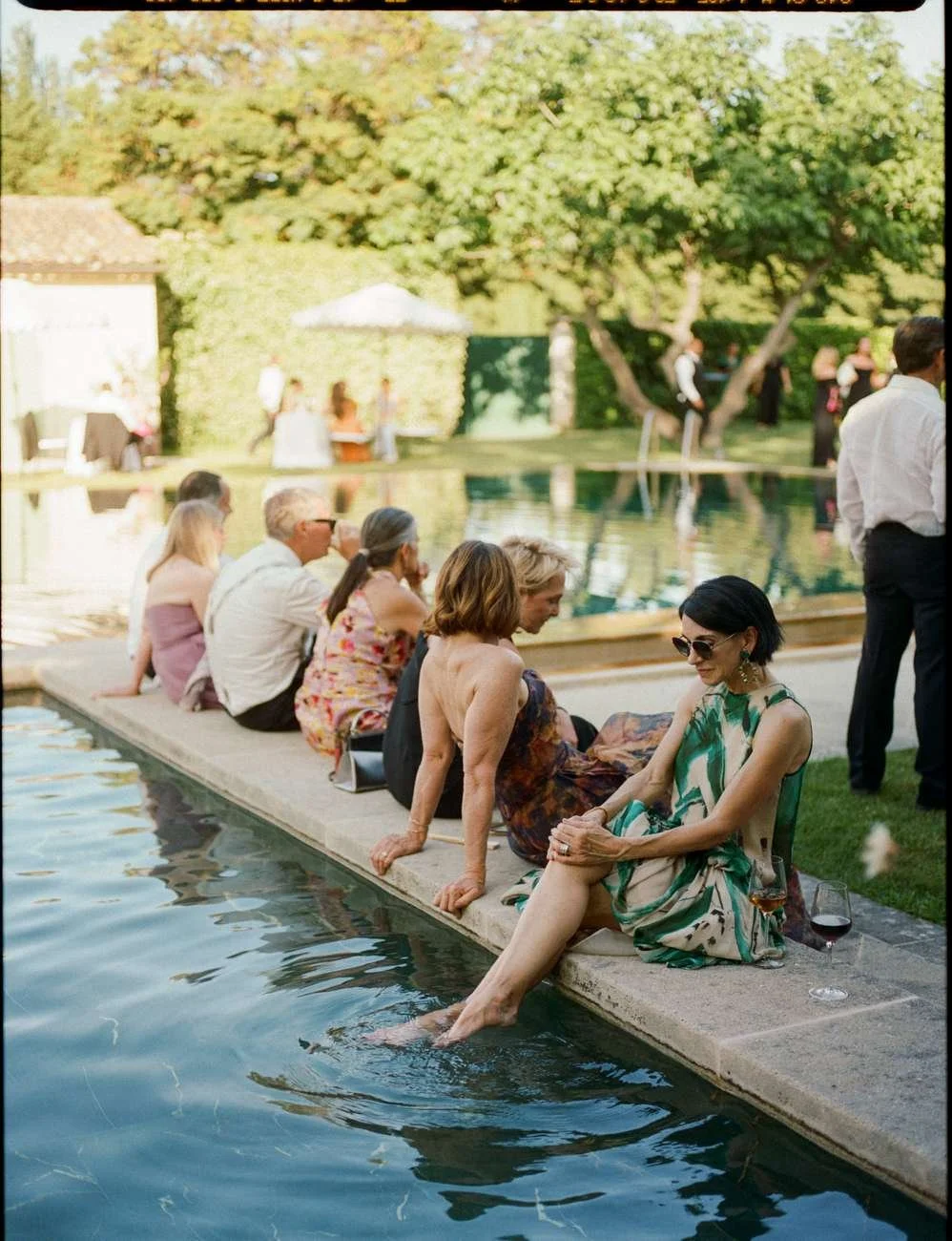 People sitting by a poolside at an outdoor gathering, with trees and other guests in the background.