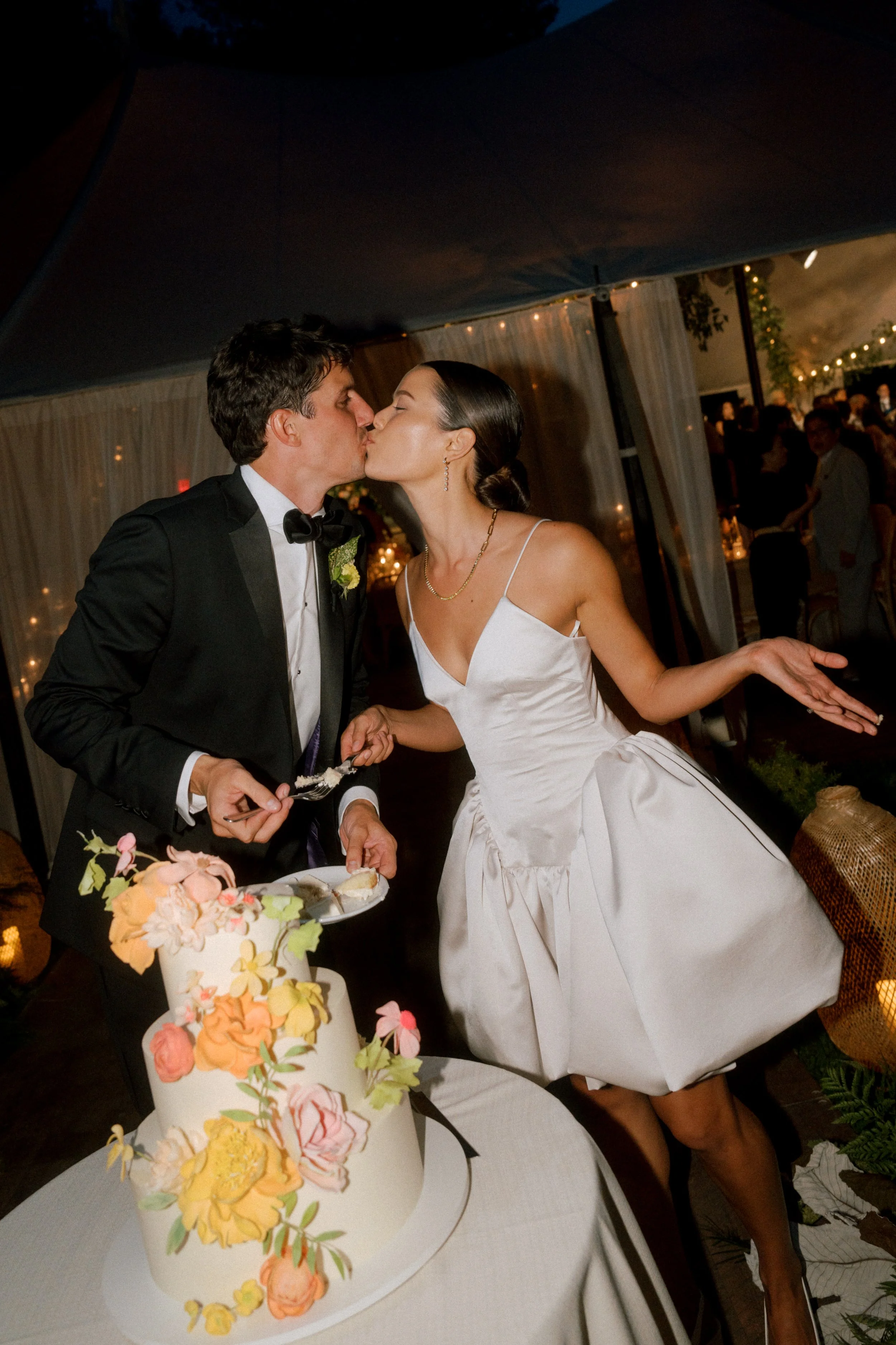 A bride and groom kiss at a wedding reception next to a decorated wedding cake.