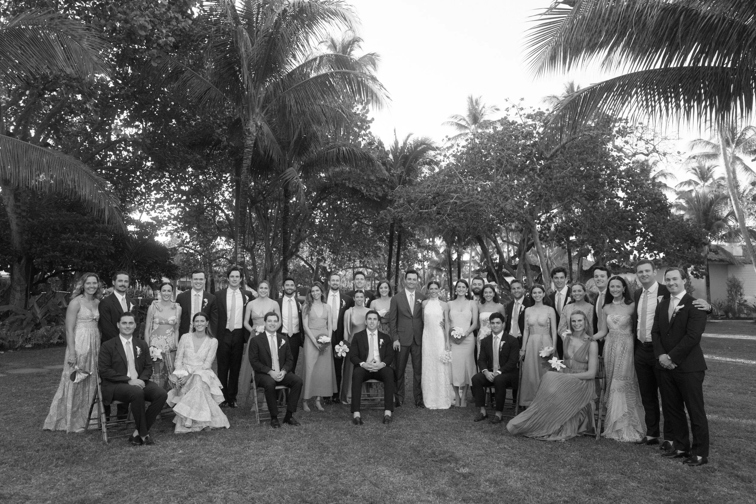 A group of people dressed in formal wedding attire, gathered outdoors in front of lush tropical trees.