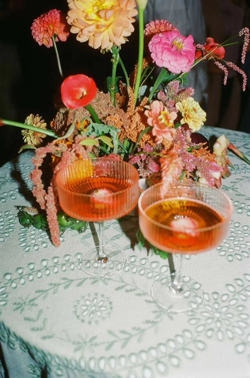 A floral centerpiece placed on a white embroidered tablecloth. Two pink cocktails in coupe glasses sit in front of the flowers.