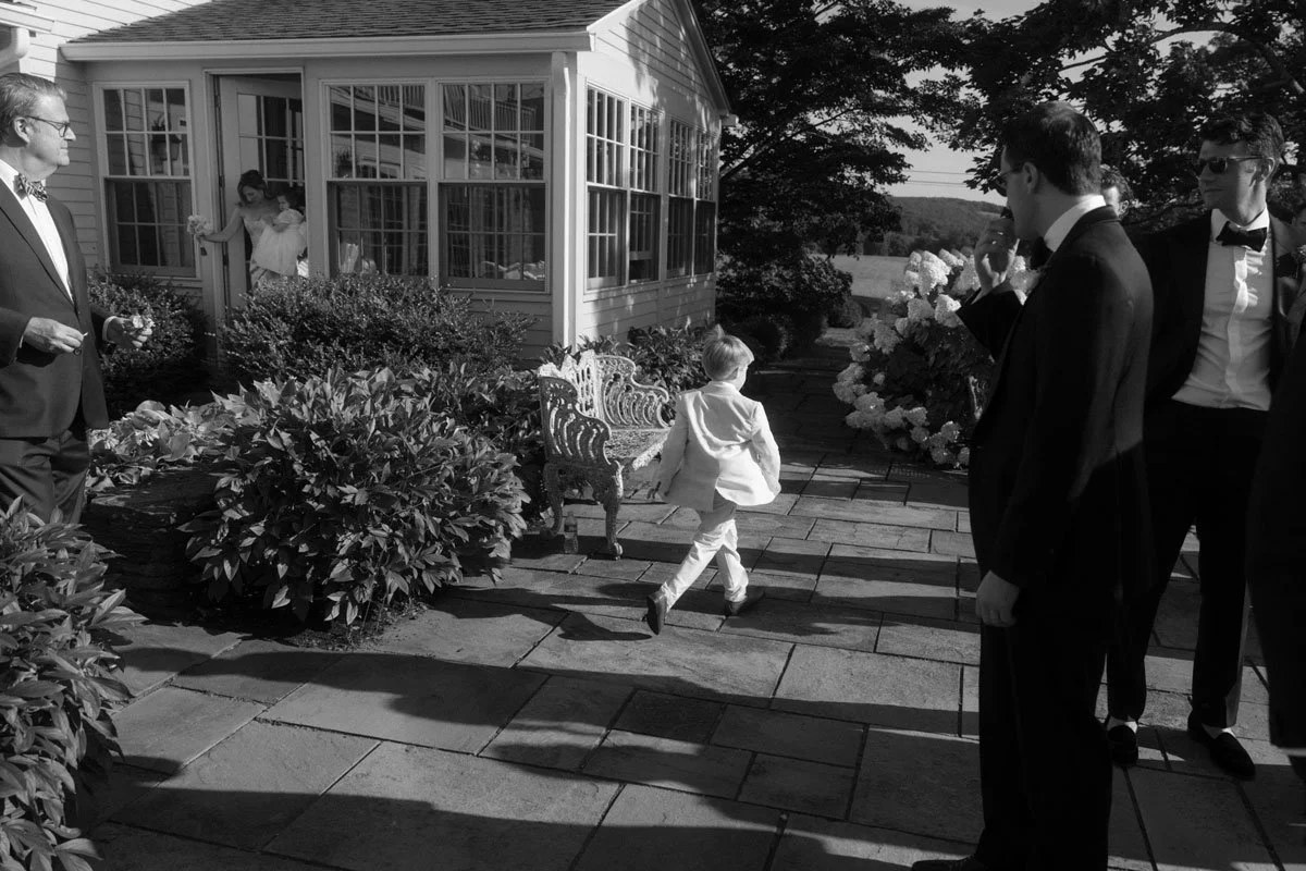 A young boy in a suit running on a paved garden path toward a group of men in tuxedos.