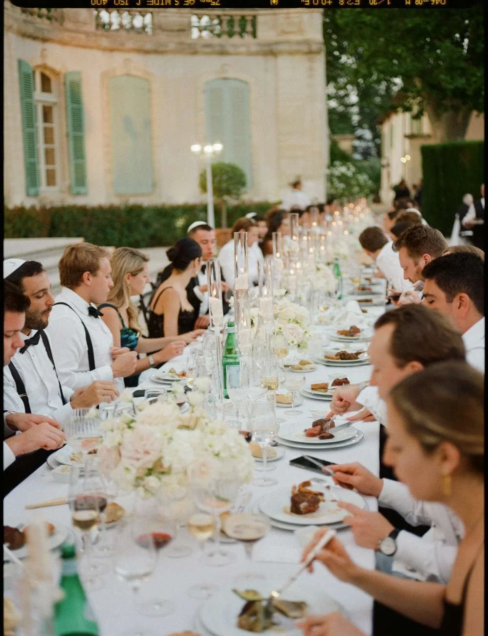 A large outdoor dinner event with many guests seated at a long table decorated with flowers and candles.