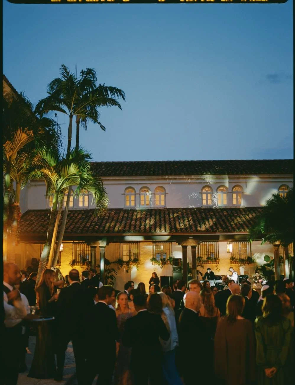 An outdoor evening event with a crowd of people dressed in formal attire, with musicians performing on a stage.