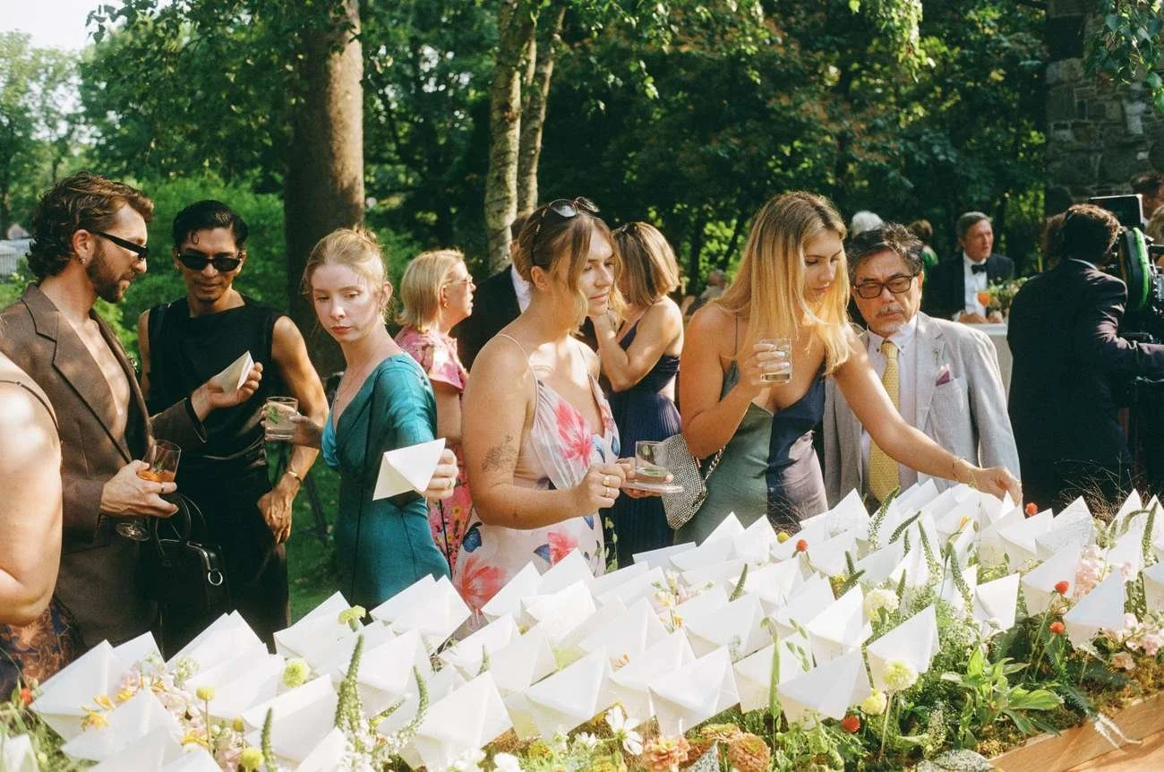 People dressed in formal attire gather outdoors around a table with white folded napkins and floral arrangements.