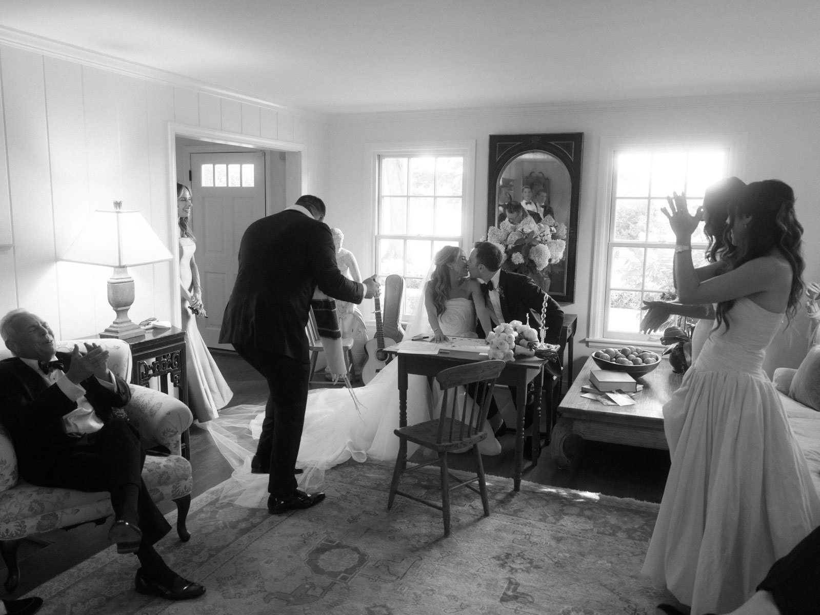 A black-and-white photo of a wedding celebration, showing a couple seated at a table sharing a kiss, surrounded by friends and family.