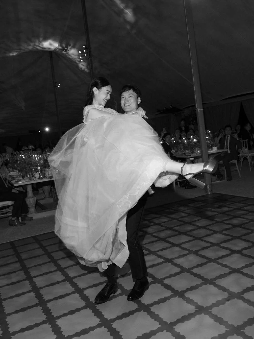 A black-and-white photo of a couple dancing at their wedding reception, the bride being carried by the groom.