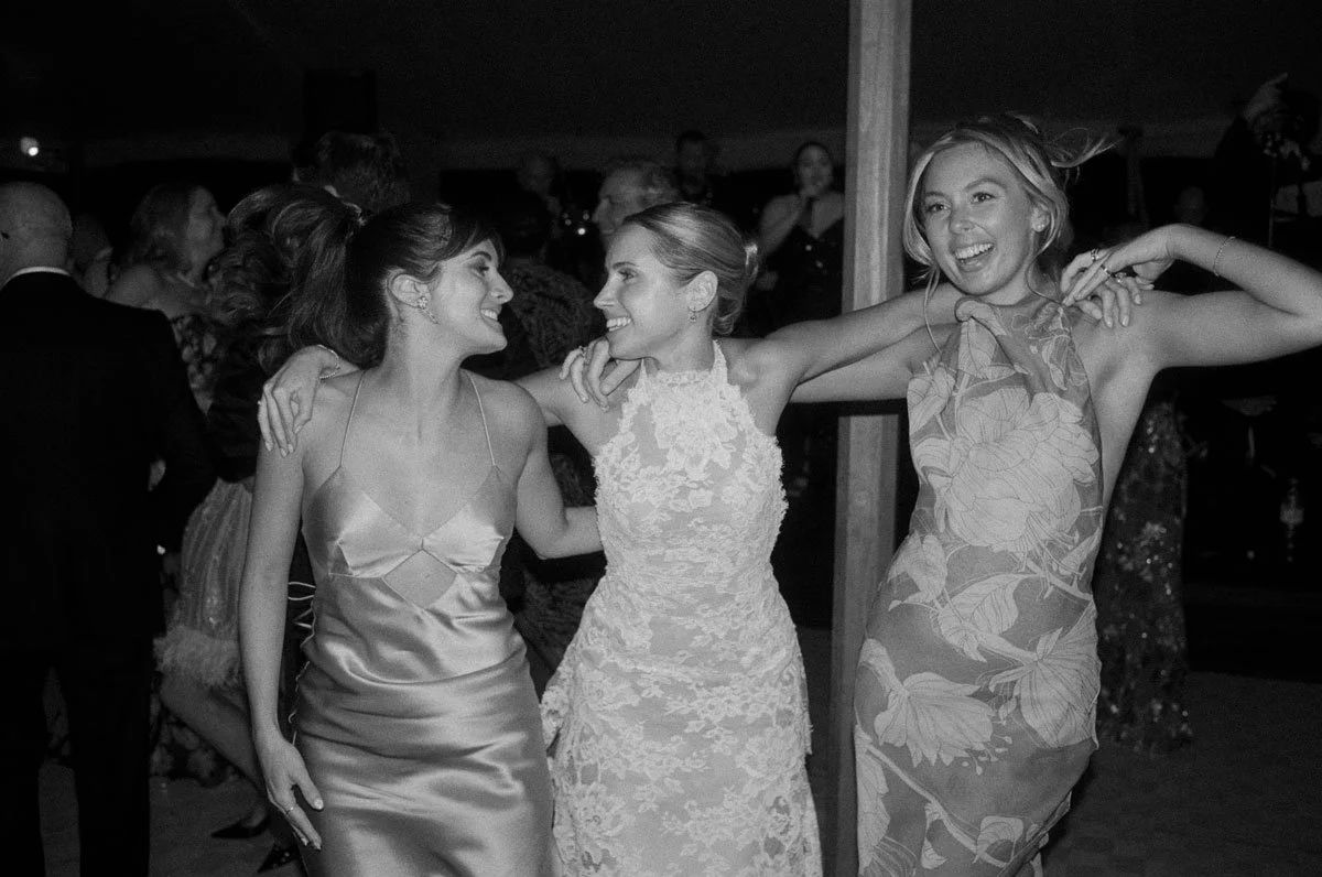 Three women in elegant dresses dancing and smiling at a celebration or wedding reception.