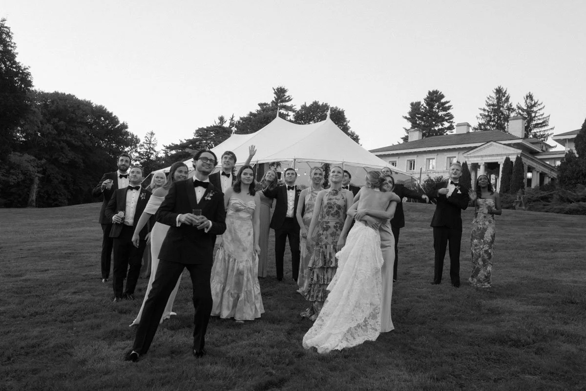 Group of people in wedding attire standing on a lawn with a large tent and house in the background.